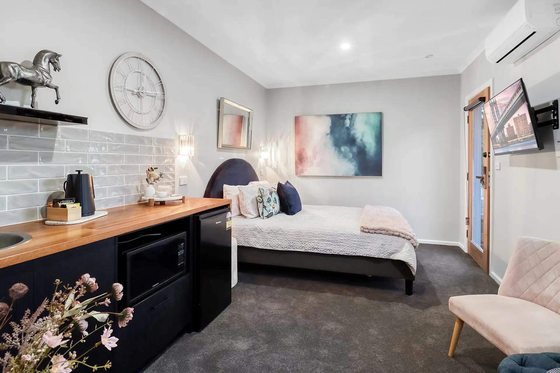 A modern hotel room features a bed, mini fridge, TV, and a plush pink chair.