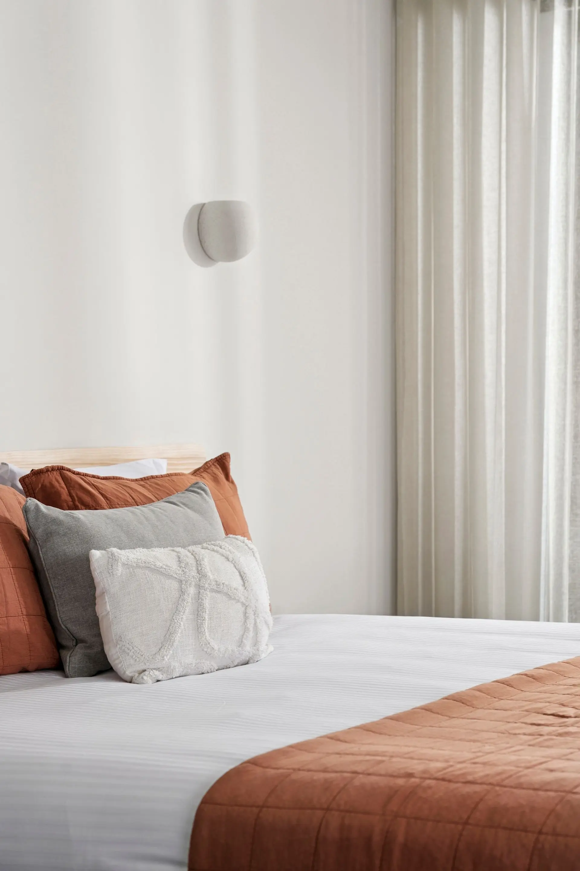 A neatly made bed with rust and gray pillows is shown, next to a cream-colored curtain.