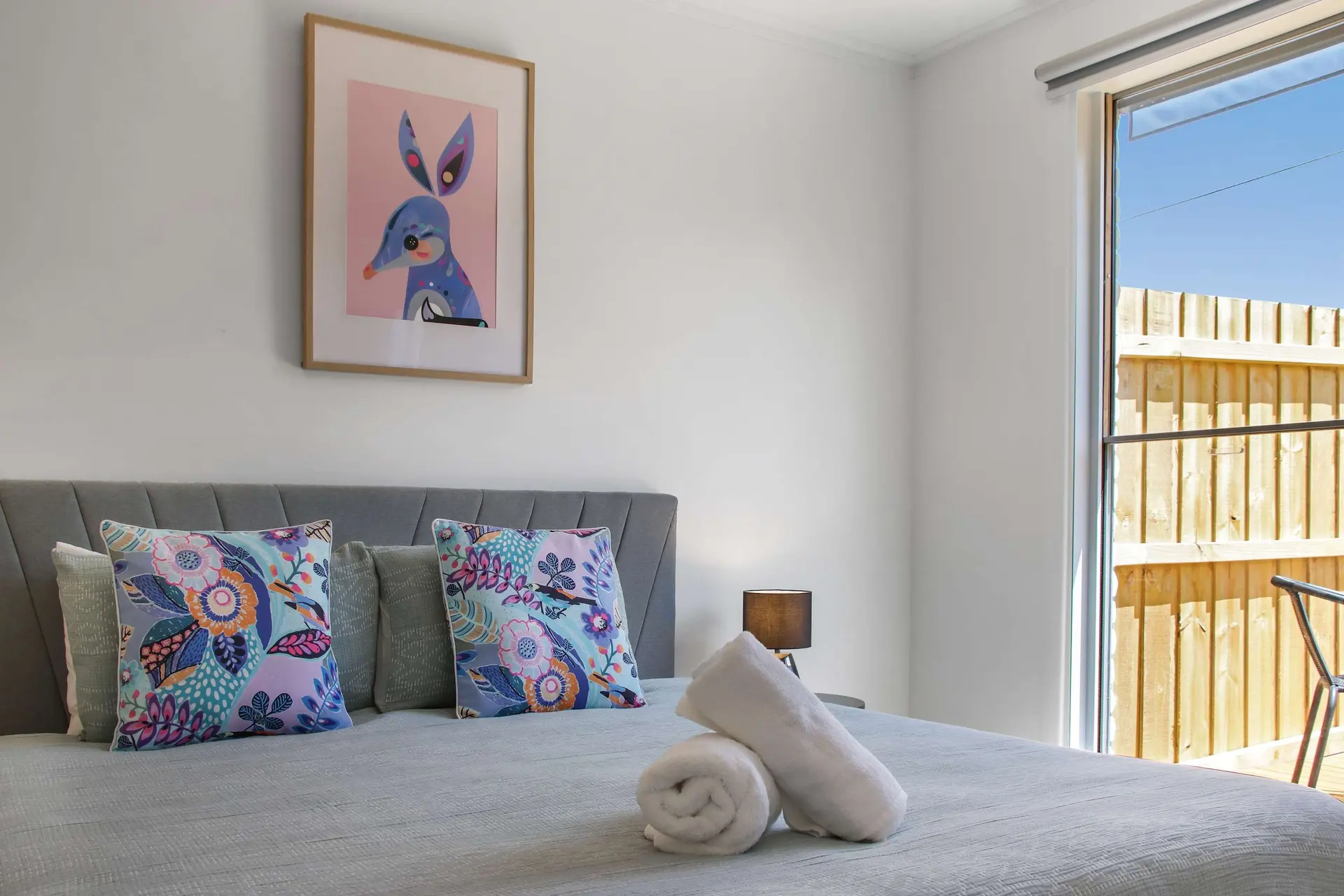 A bedroom with a gray headboard, colorful pillows, and a framed picture of a blue animal.