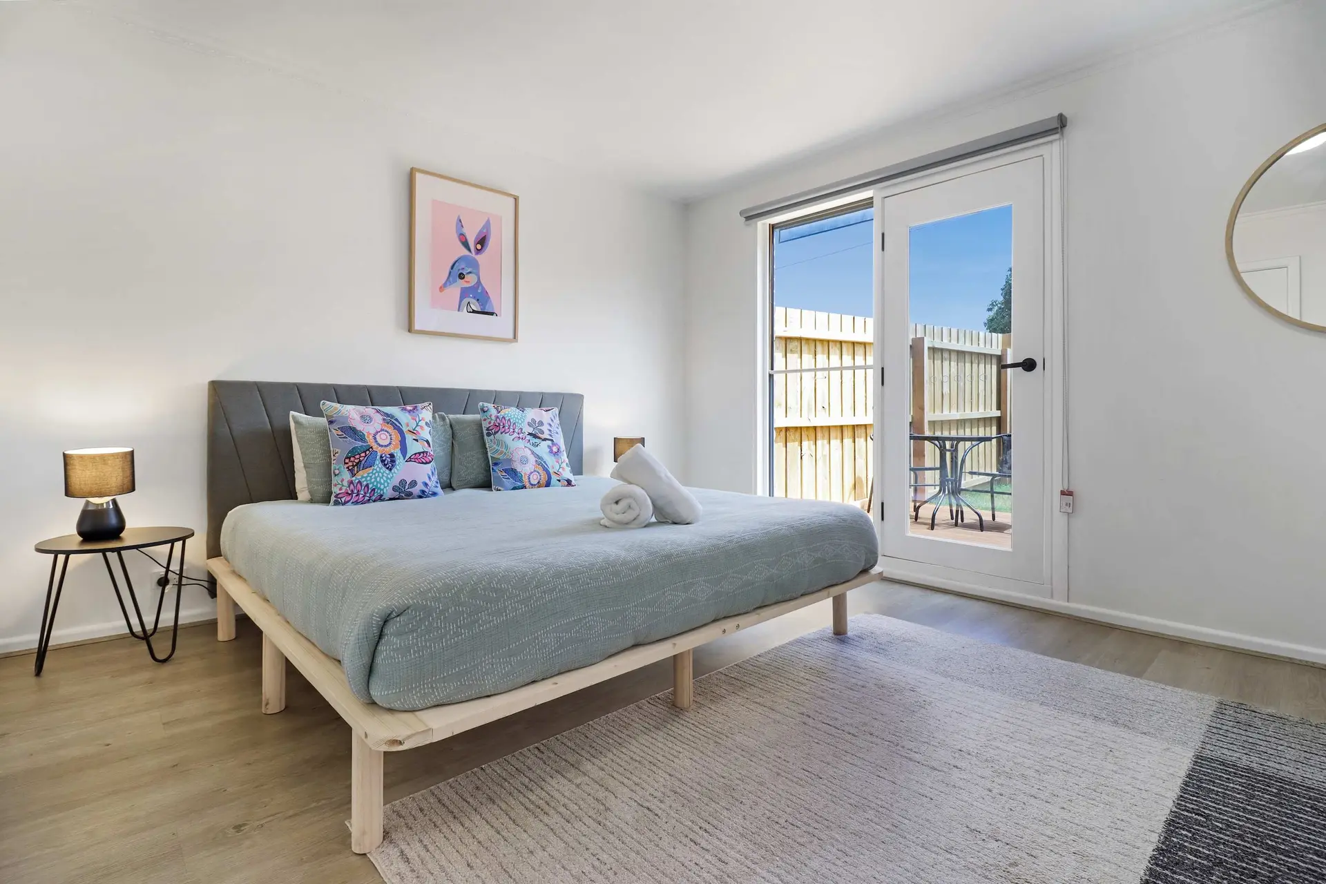A bright bedroom with a light wood floor, a large bed with patterned pillows, and a sliding glass do