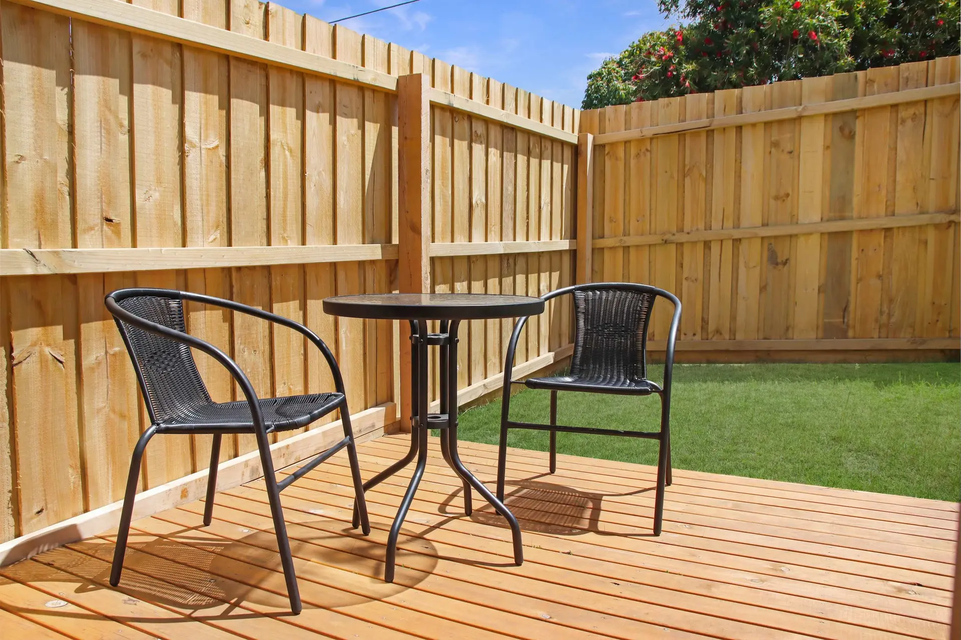A wooden deck with a table and two chairs, surrounded by a wooden fence, with green grass and a tree