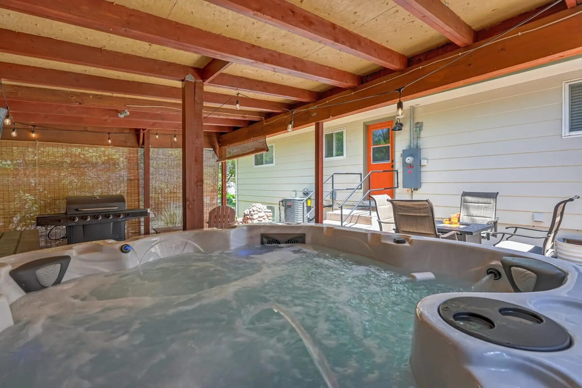 Bubbling hot tub on a covered patio with a grill, outdoor dining, and a house in the background.