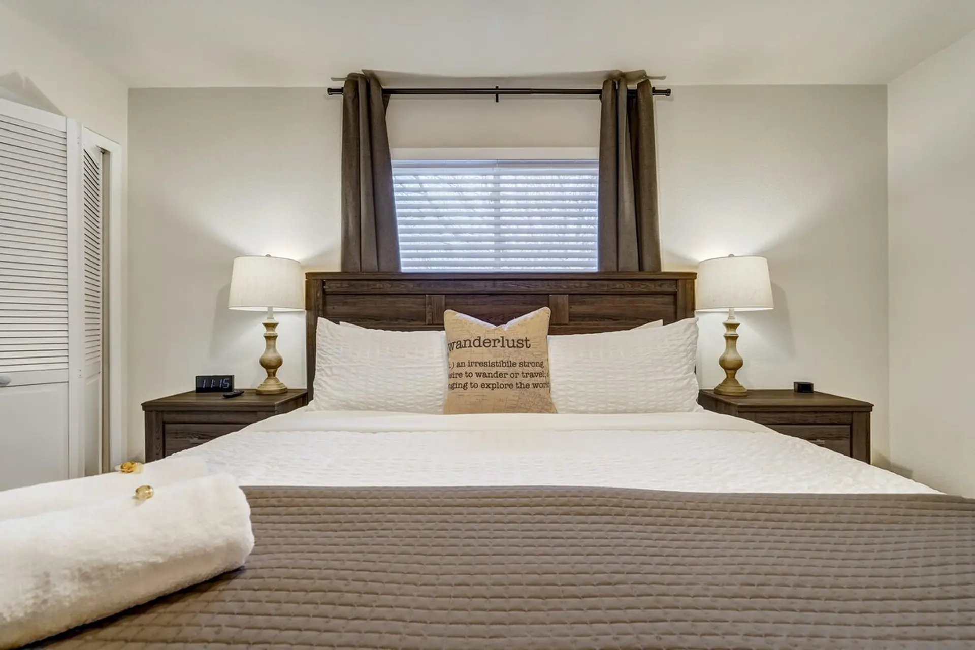 A bedroom featuring a large bed with white bedding, 'wanderlust' pillow, and two nightstands with la