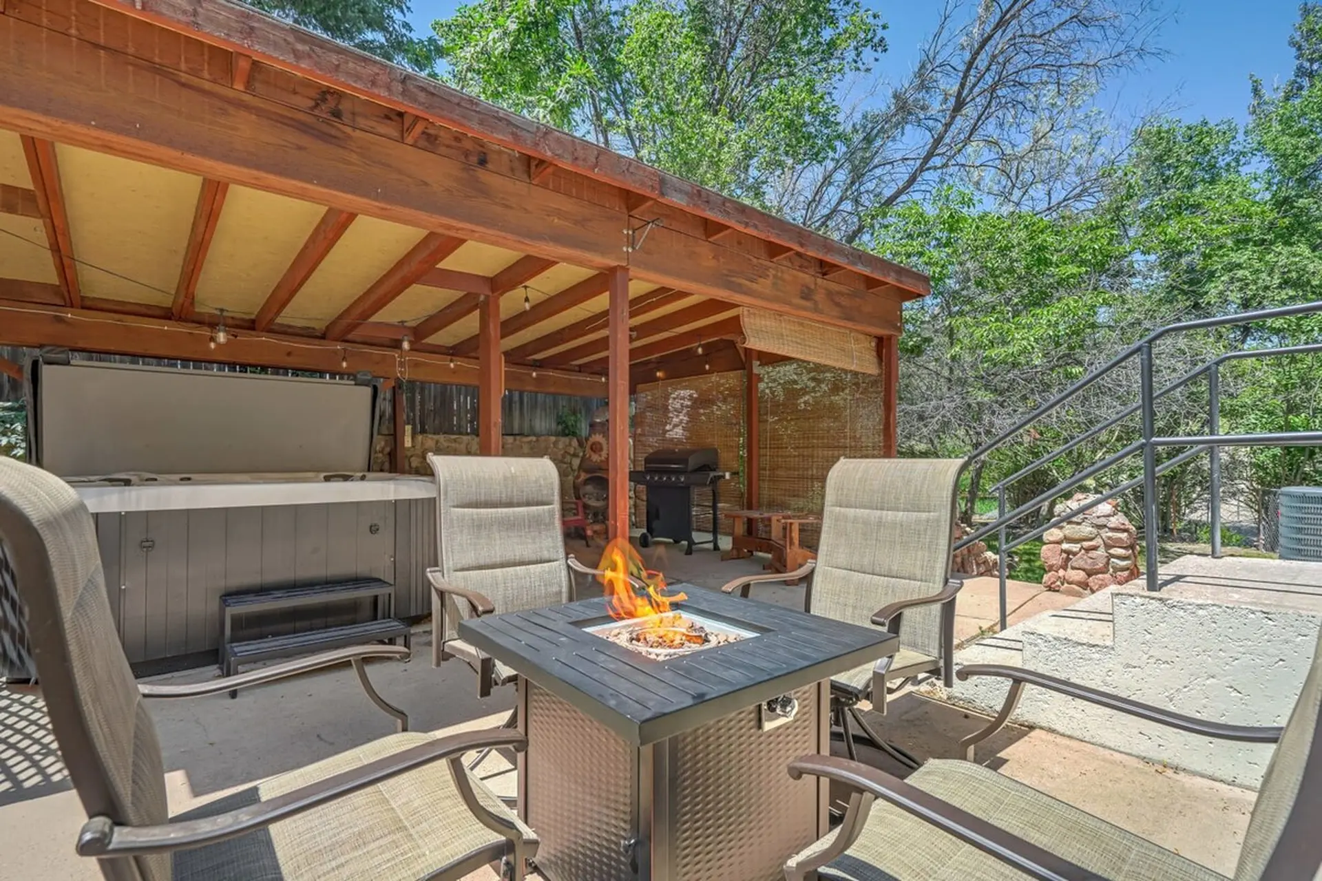 Outdoor patio under a wooden pergola with a hot tub, lighted fire pit, BBQ grill, and chairs.