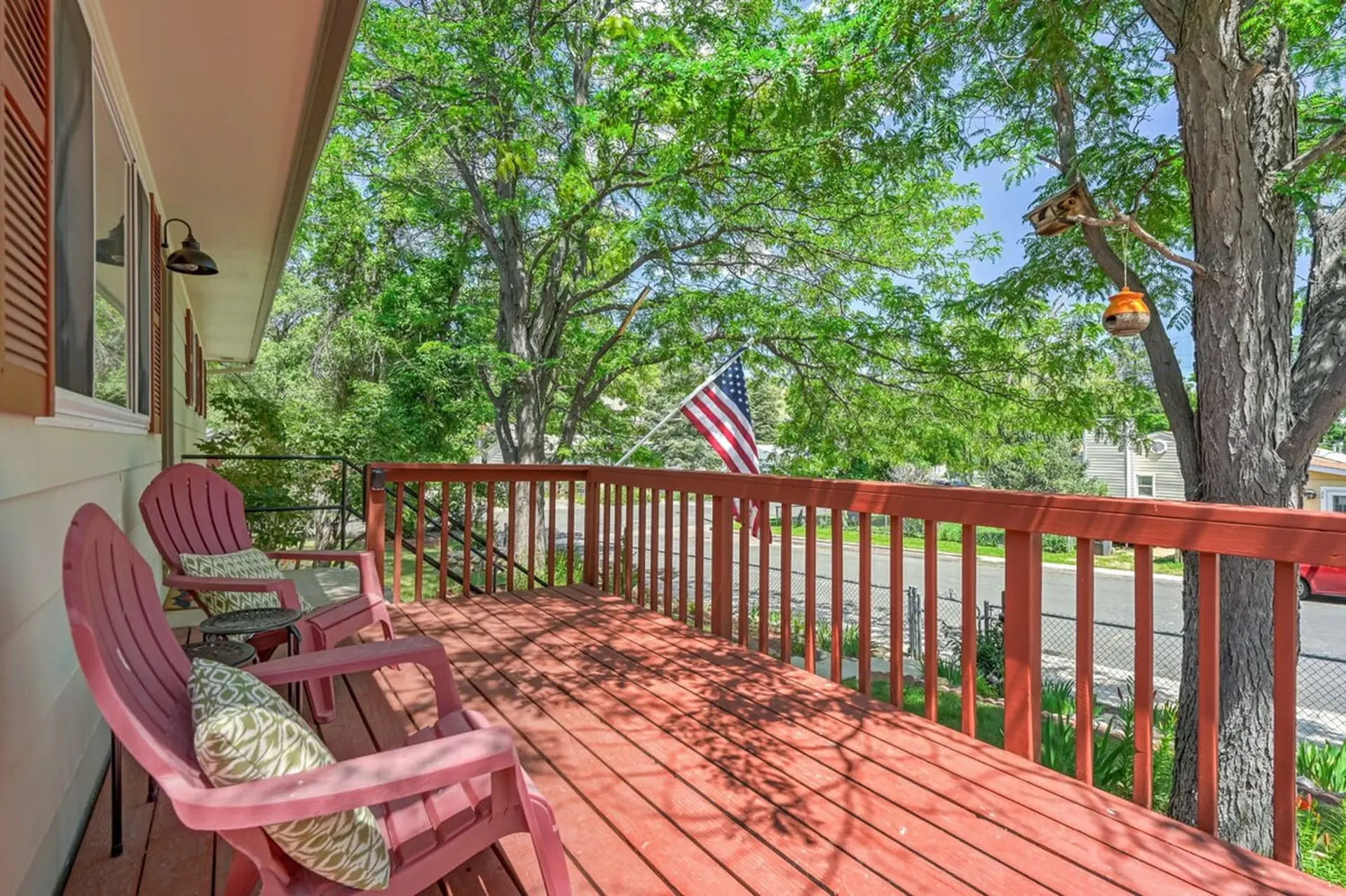 A red wooden porch with two pink chairs, a US flag, and lush green trees in the background.