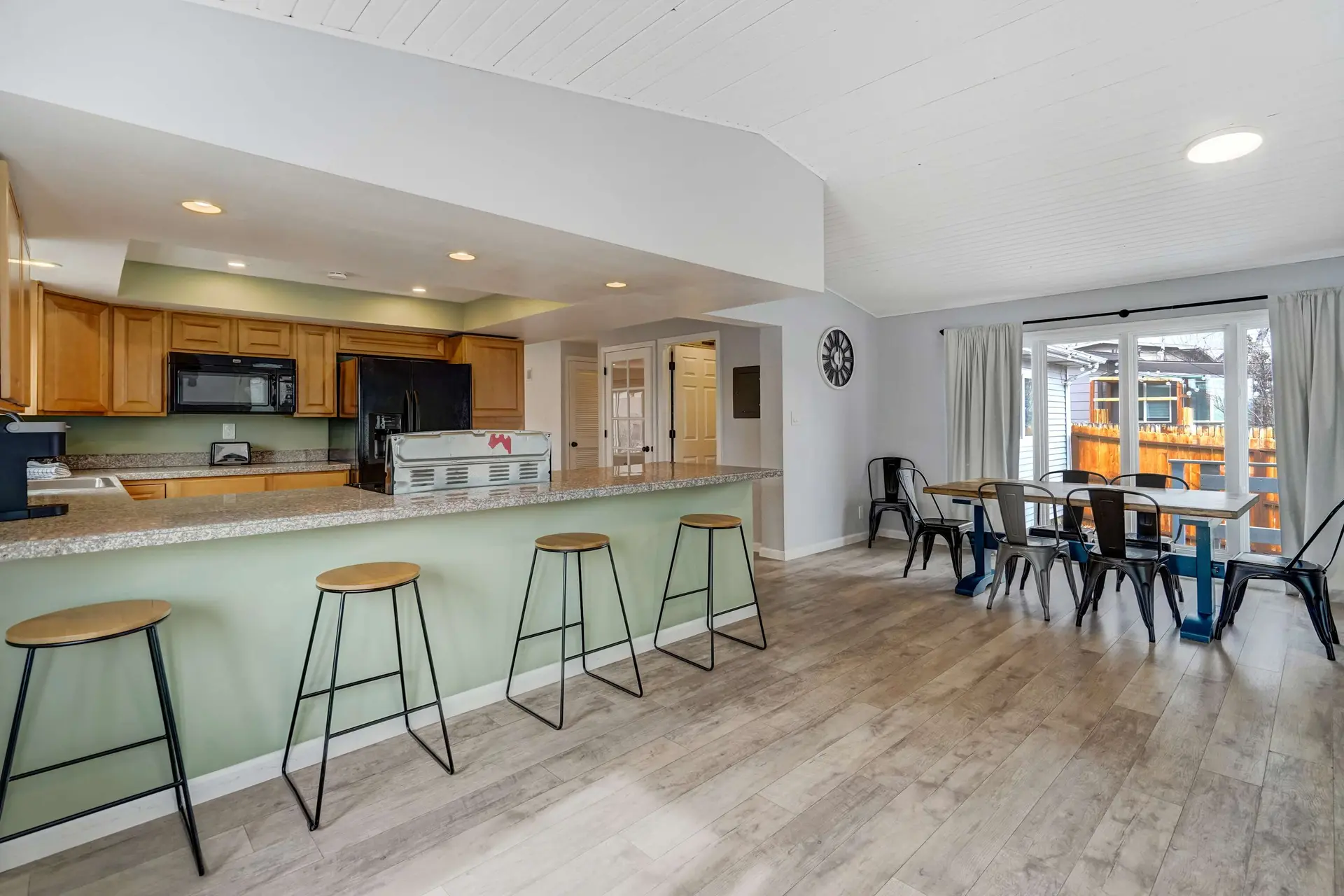 Bright open kitchen and dining area with wood cabinets, green bar, and modern table with metal chair