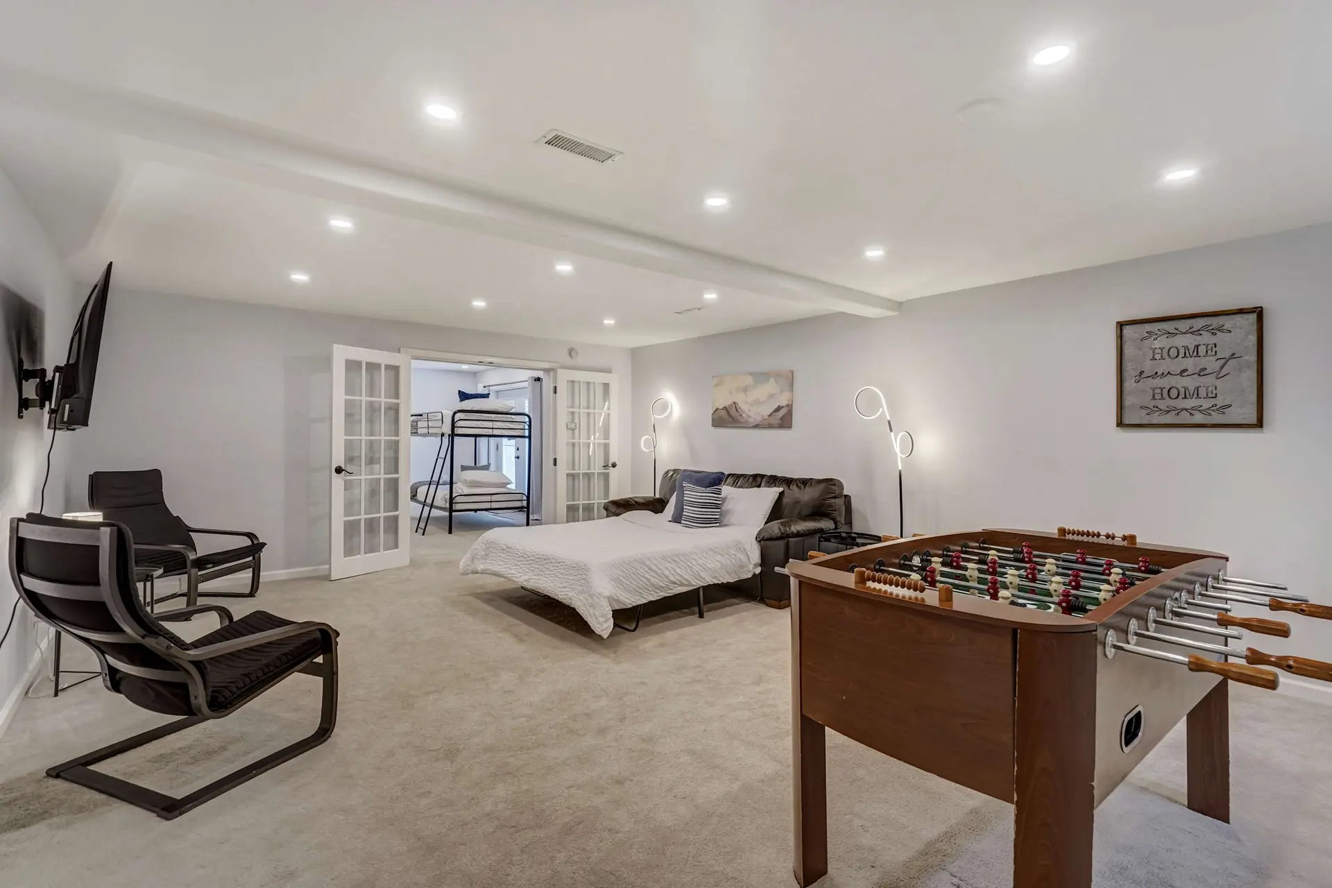 A bright basement rec room with a foosball table, a sofa bed, a TV area, and a bunk bed room.