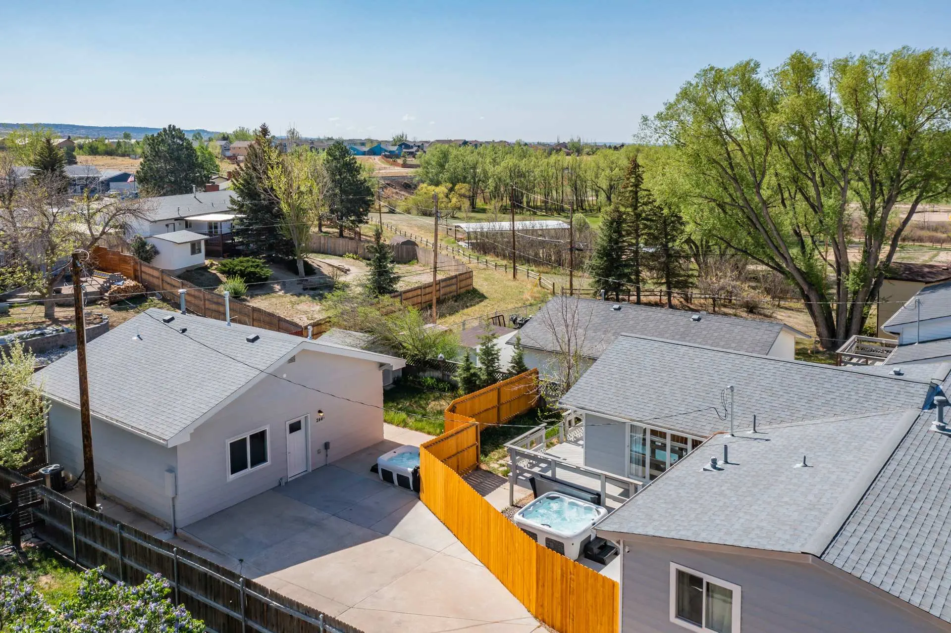 Aerial view of houses with gray roofs, two hot tubs, a long wooden fence, and green trees.