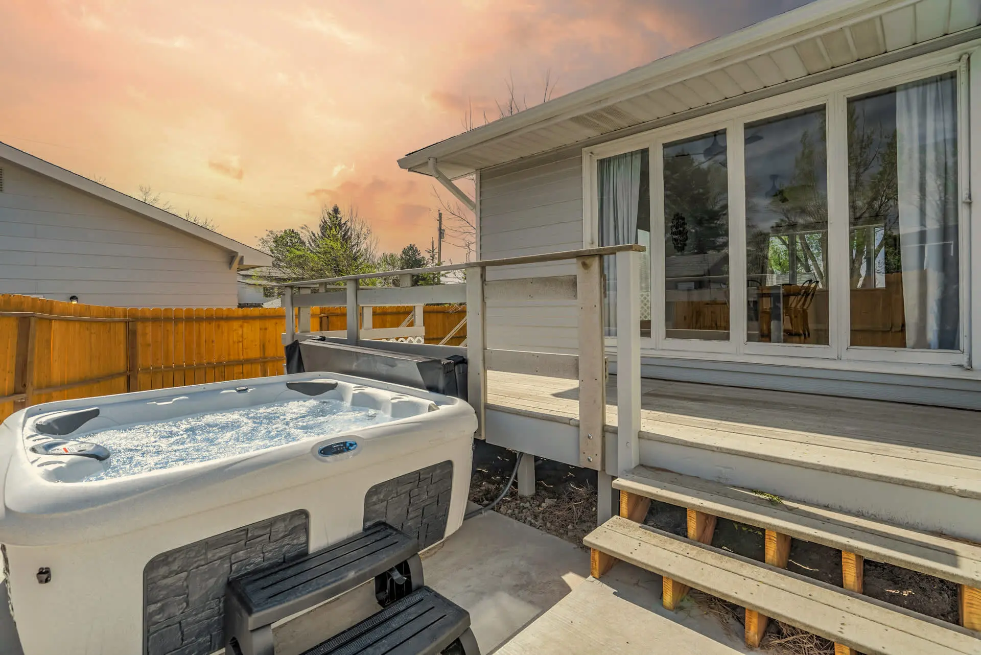 A bubbling hot tub on a concrete patio next to a wooden deck with steps, connected to a house.
