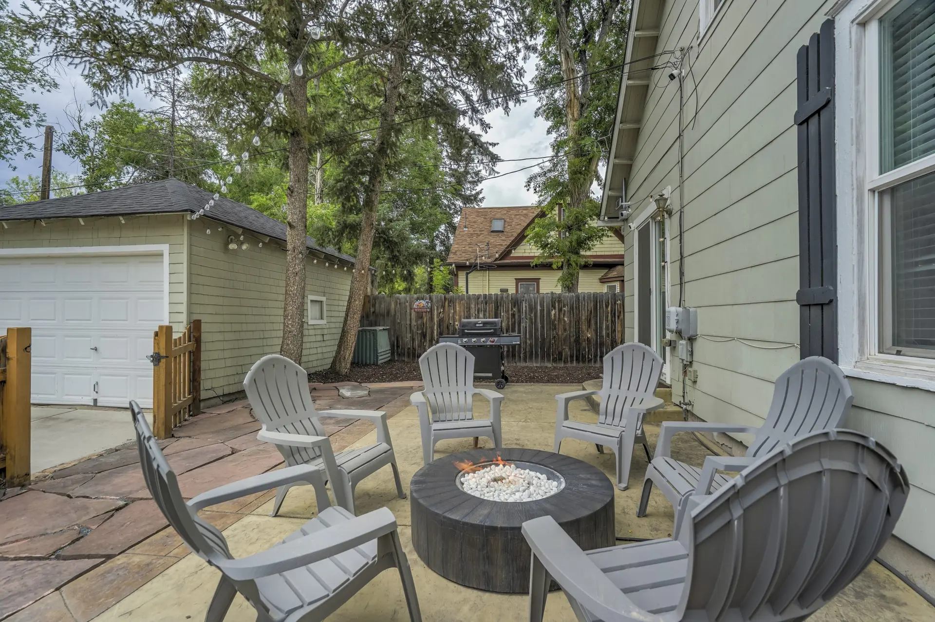 An outdoor patio with a lit fire pit, six grey Adirondack chairs, a house, garage, and trees.
