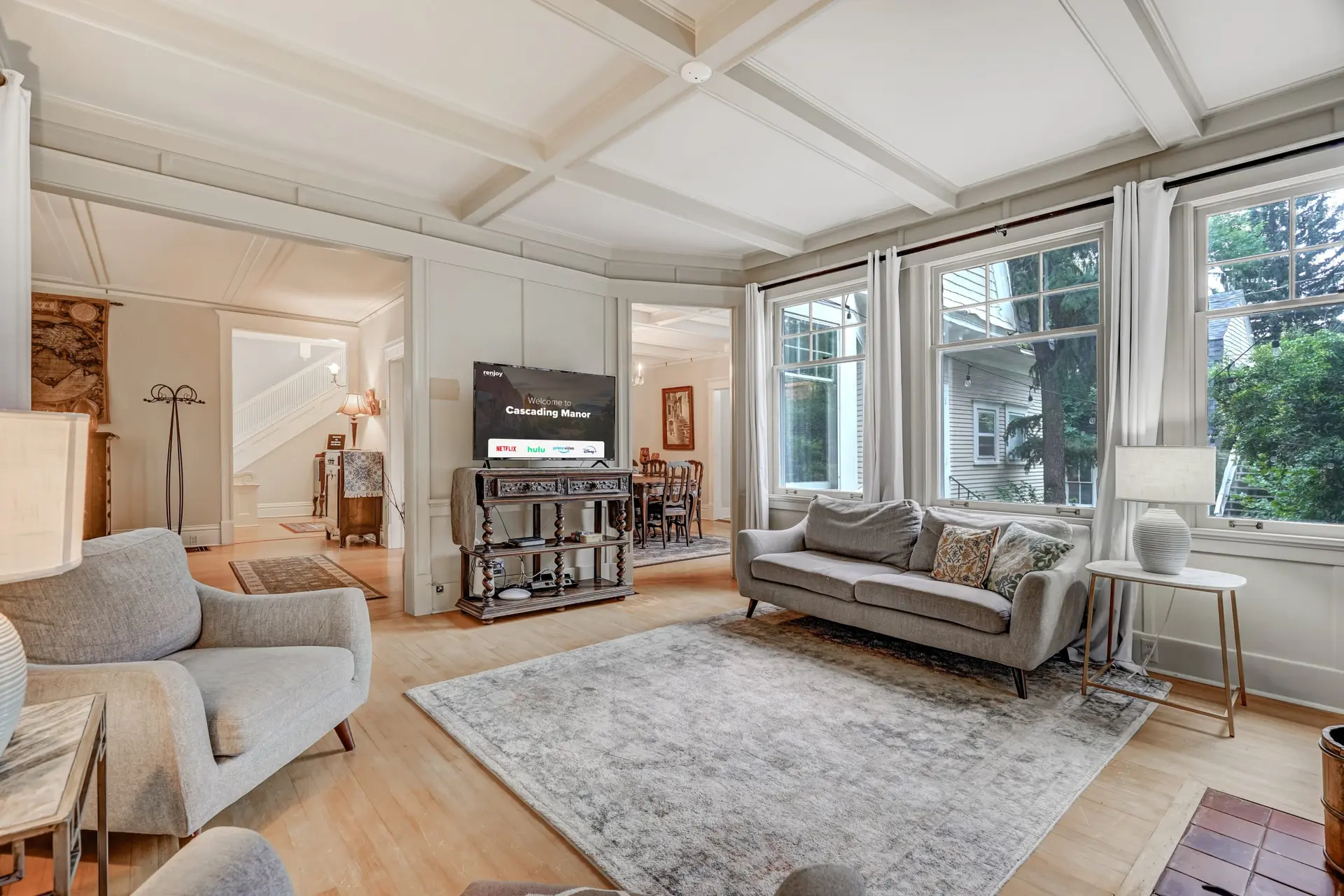 A bright living room with gray furniture, a TV on a console, a light rug, and a coffered ceiling.