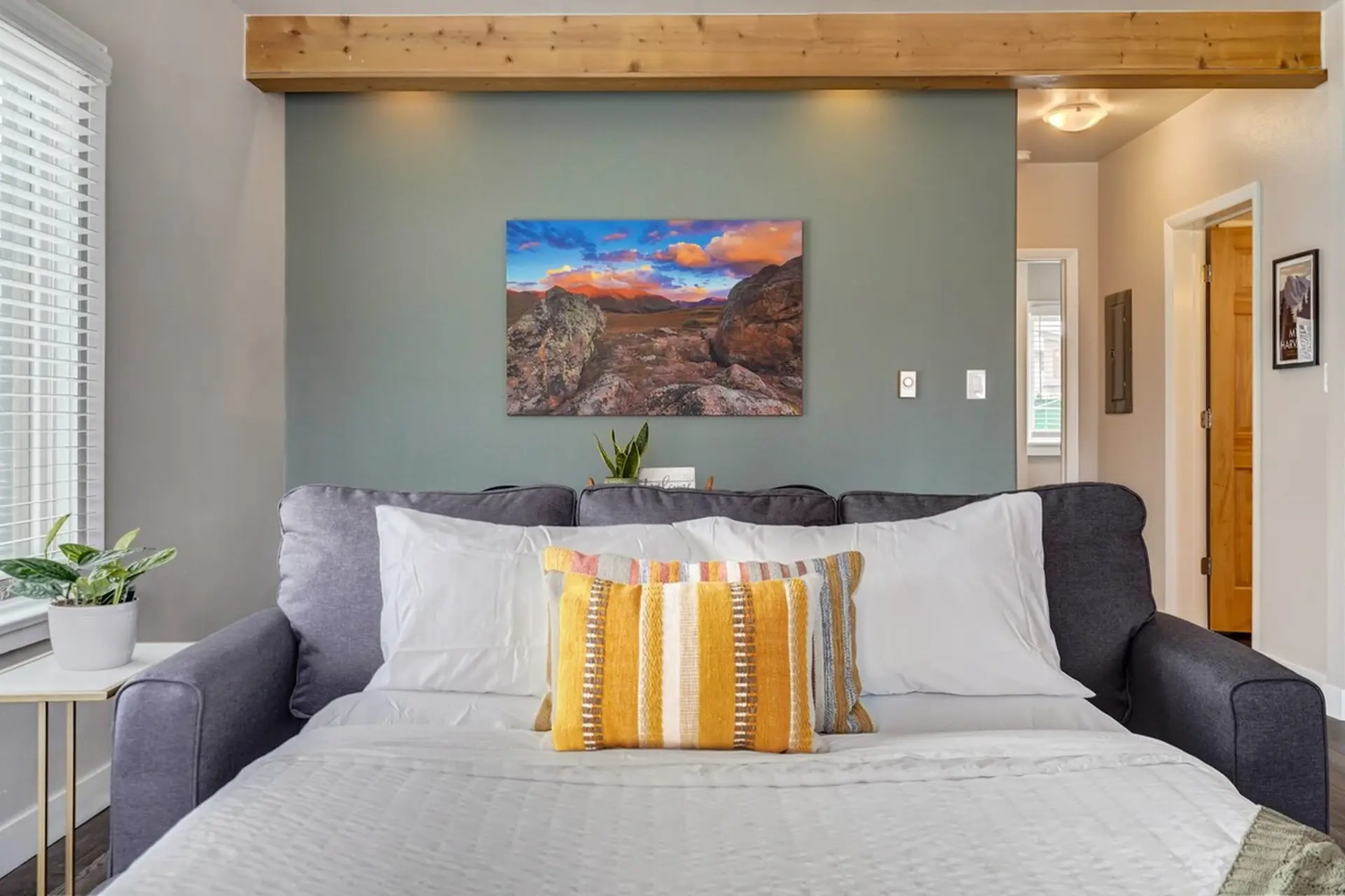 A sofa bed with white bedding and a yellow striped pillow against a sage green wall with landscape a