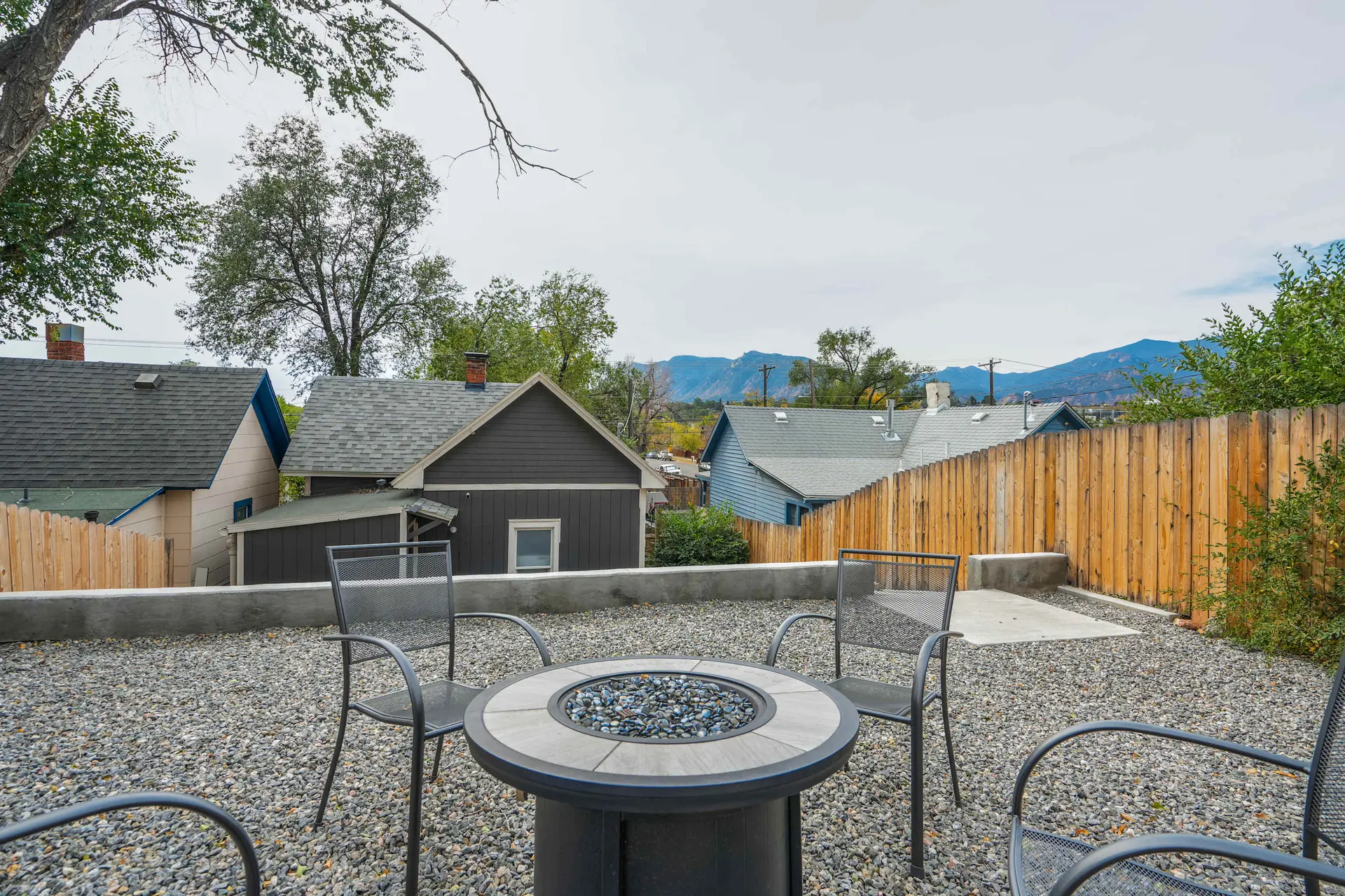 An outdoor patio with a fire pit and chairs is surrounded by houses and mountains.