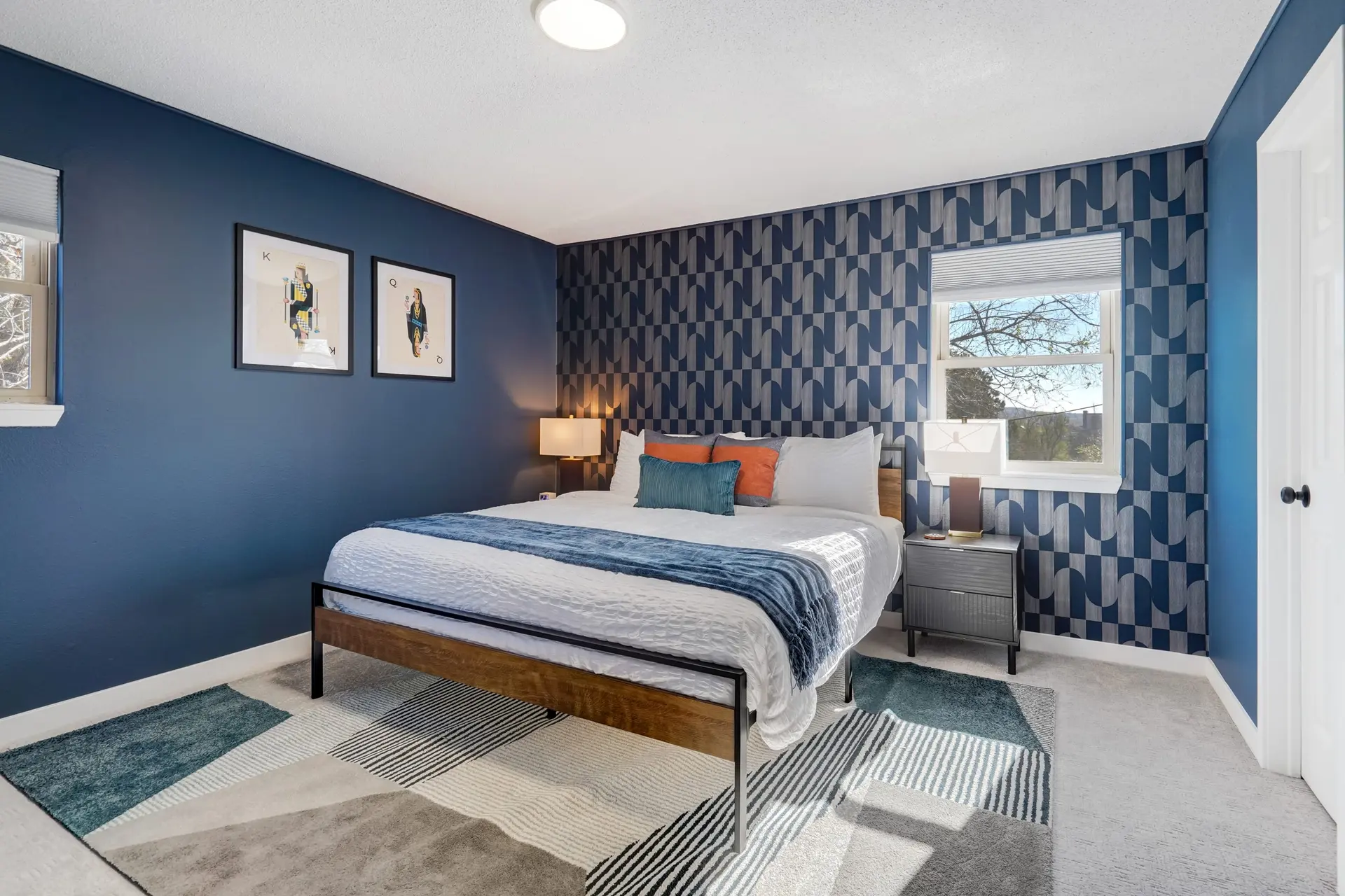 Modern blue bedroom with patterned wallpaper, a bed, geometric rug, and nightstands.
