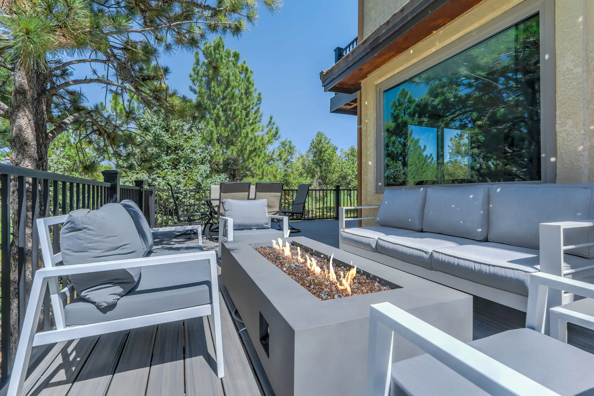 Outdoor deck with modern gray patio furniture, a lit fire pit, surrounded by trees and a blue sky.