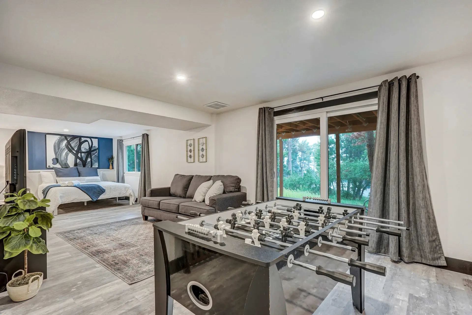 Spacious basement with a bed, sofa, foosball table, and large window overlooking a forest.