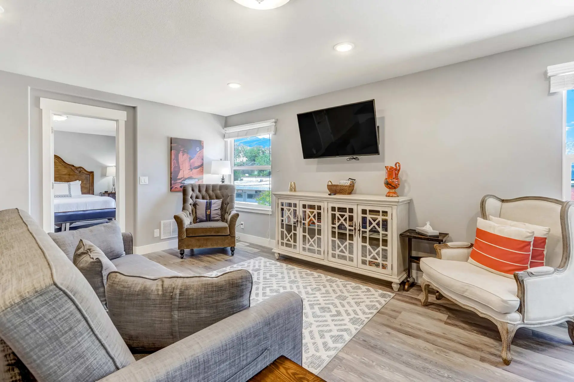 A well-lit living room with gray sofa, armchairs, TV, and a view into a bright bedroom.