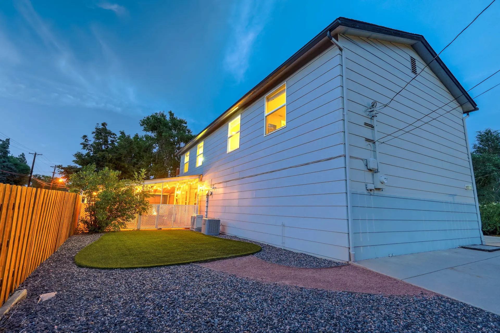 Two-story house exterior at dusk with warm light from windows/patio, gravel yard, and wooden fence.