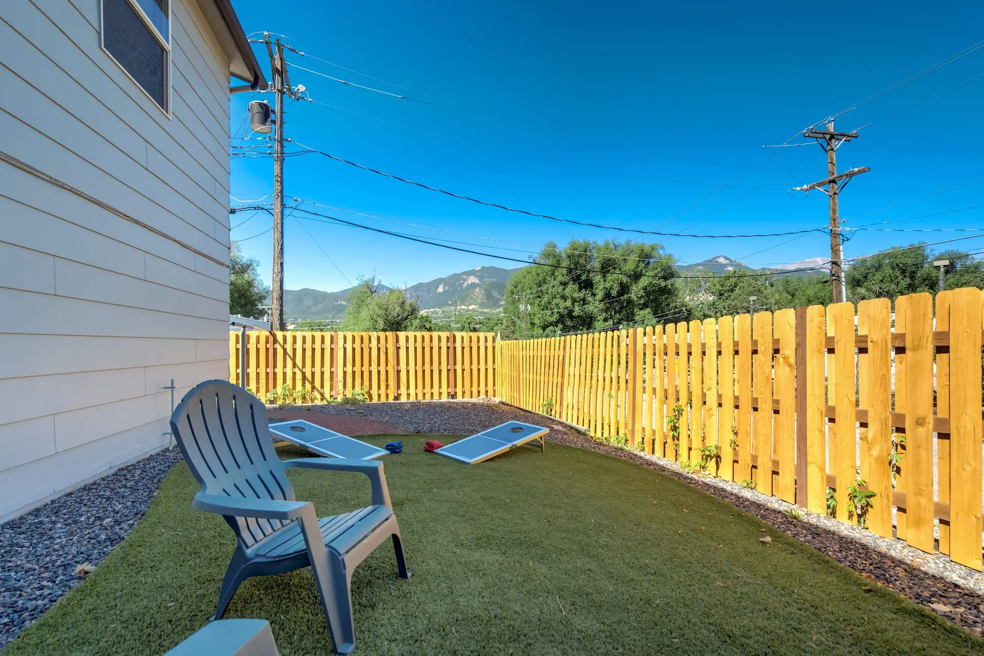 Backyard with artificial grass, cornhole boards, fence, and mountains under a blue sky.