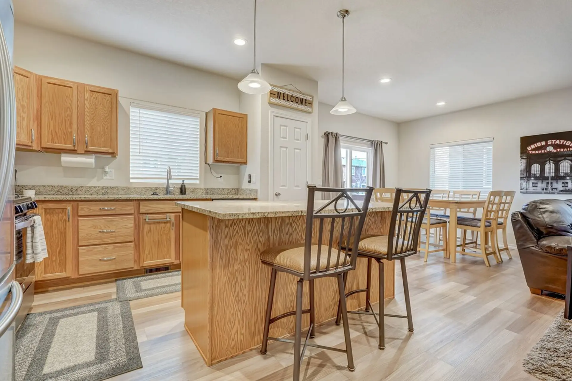 A bright, open-concept kitchen with light wood cabinets, granite island, bar stools, and dining area