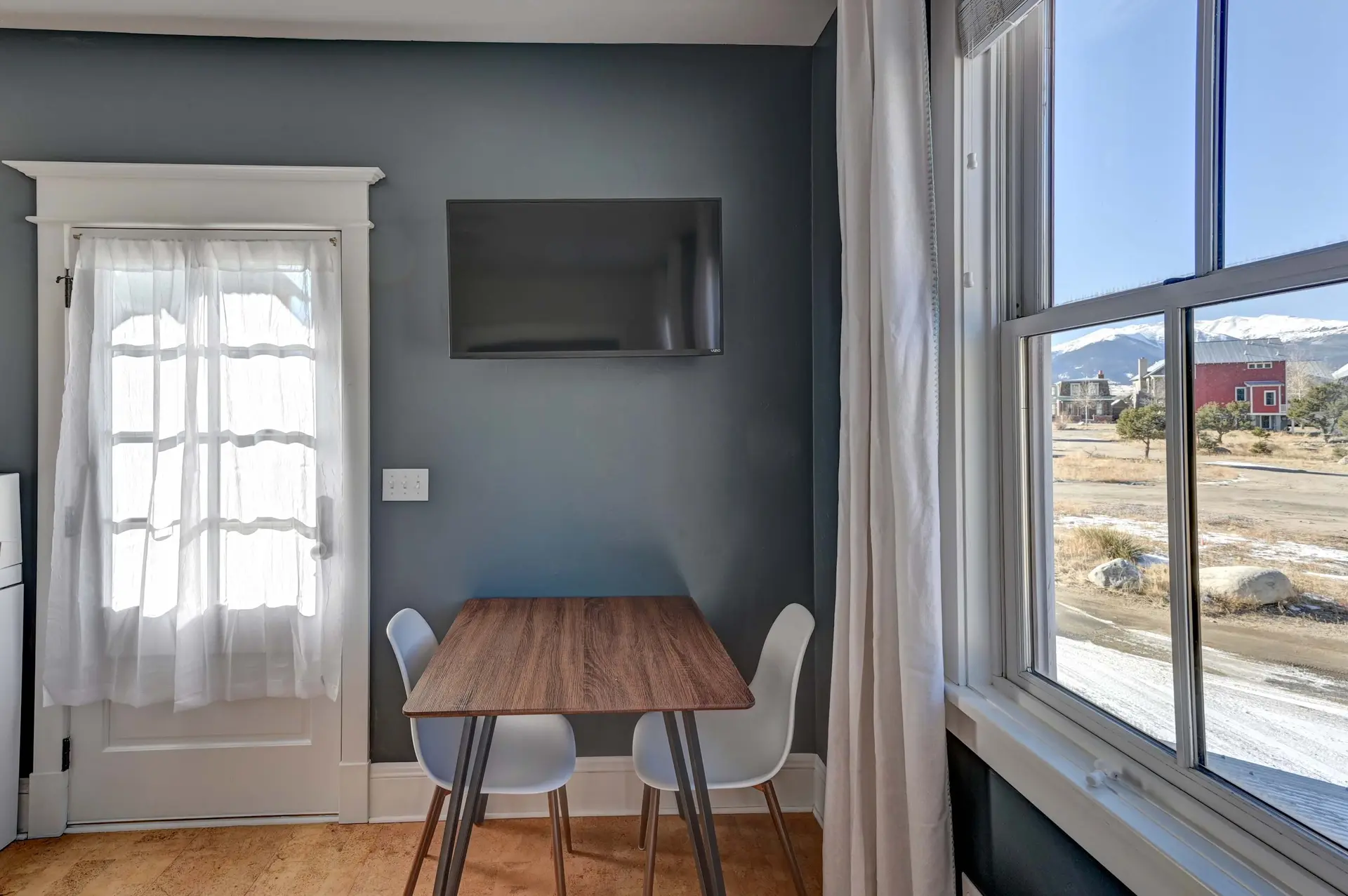 A dining table and chairs in a room with a tv on the wall and a window view.