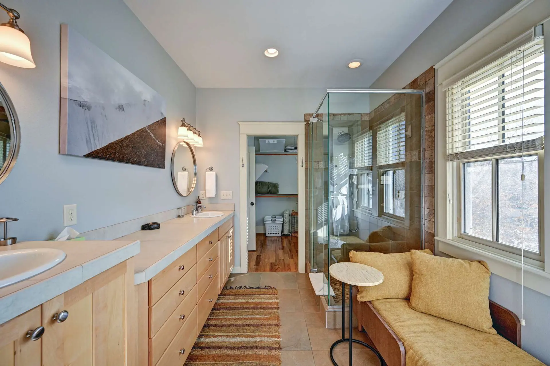 A bathroom with dual sinks, a glass shower, and a window seat.