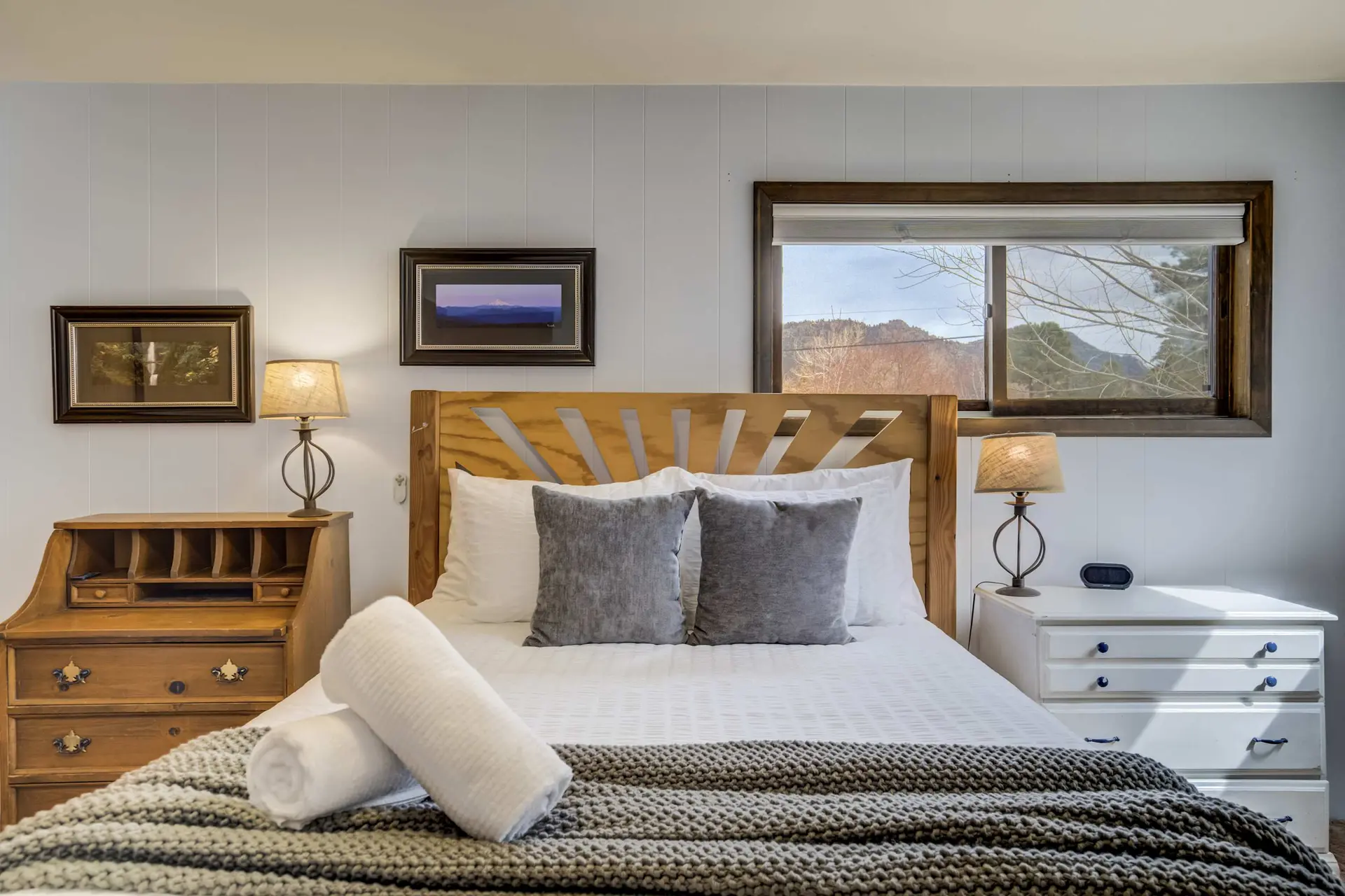 Bright bedroom with a bed, white and gray bedding, two lamps, wood and white dressers, and window.