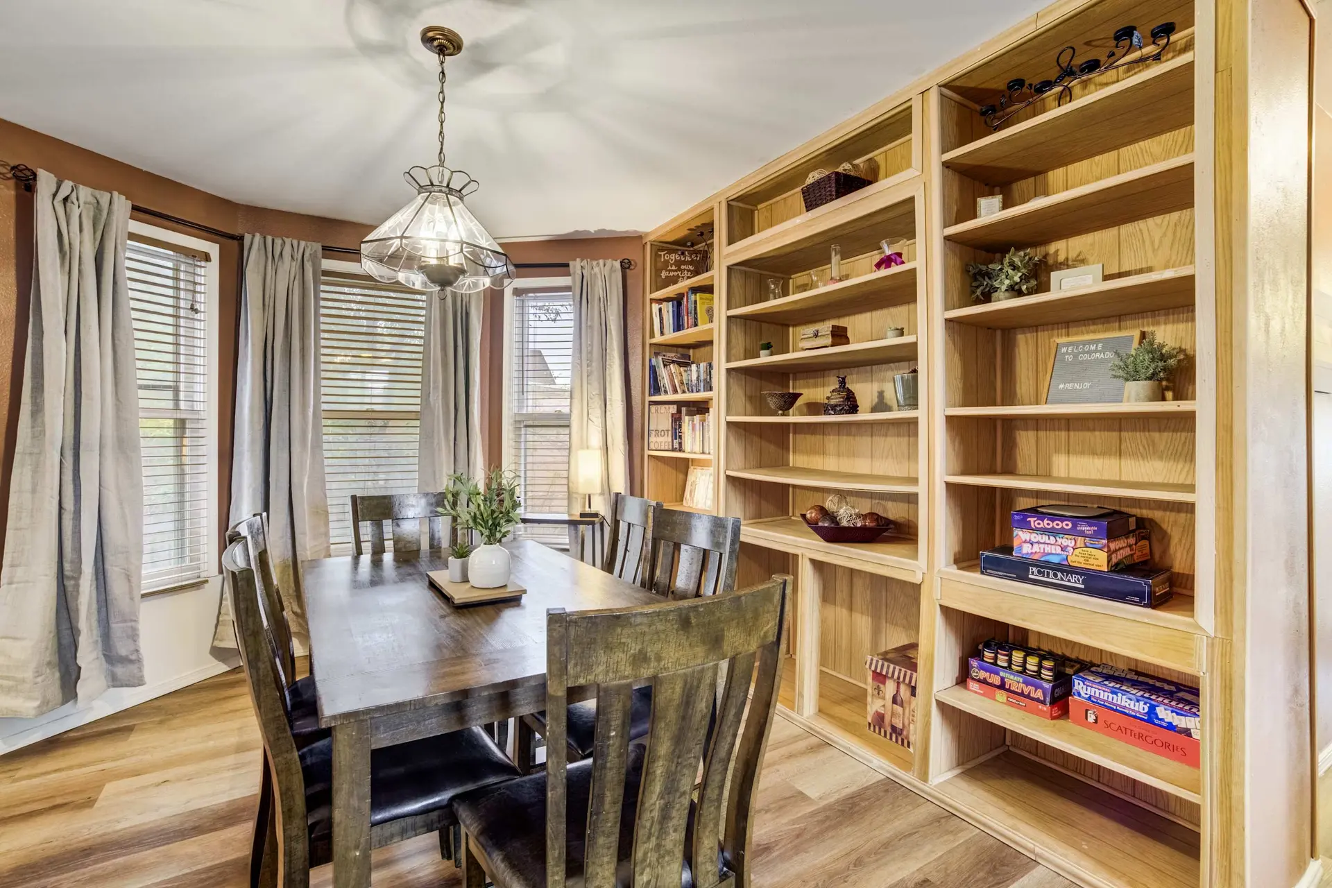 A dining table with chairs is in front of a large bookshelf filled with books and games.