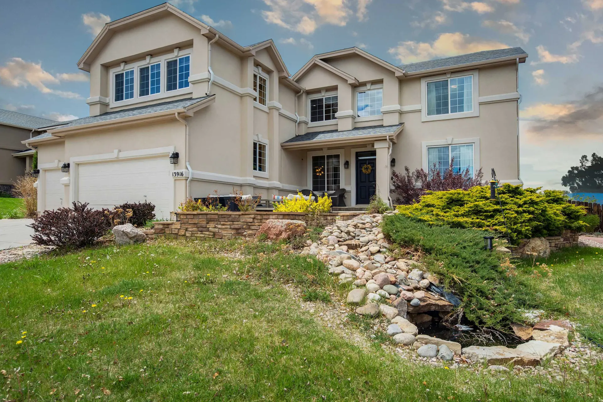 Large, tan two-story house with green lawn, varied landscaping, and a dry rock stream.