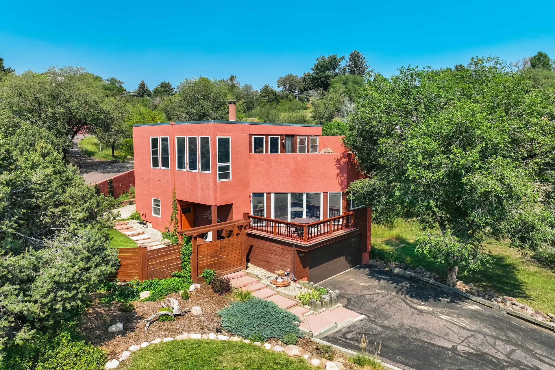 Aerial view of a red-orange modern house with a balcony, driveway, green trees, and a blue sky.