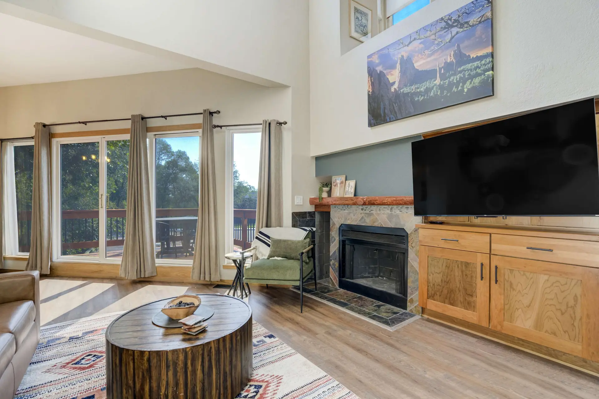 A cozy living room with a fireplace, TV, large mountain painting, and big windows overlooking trees.