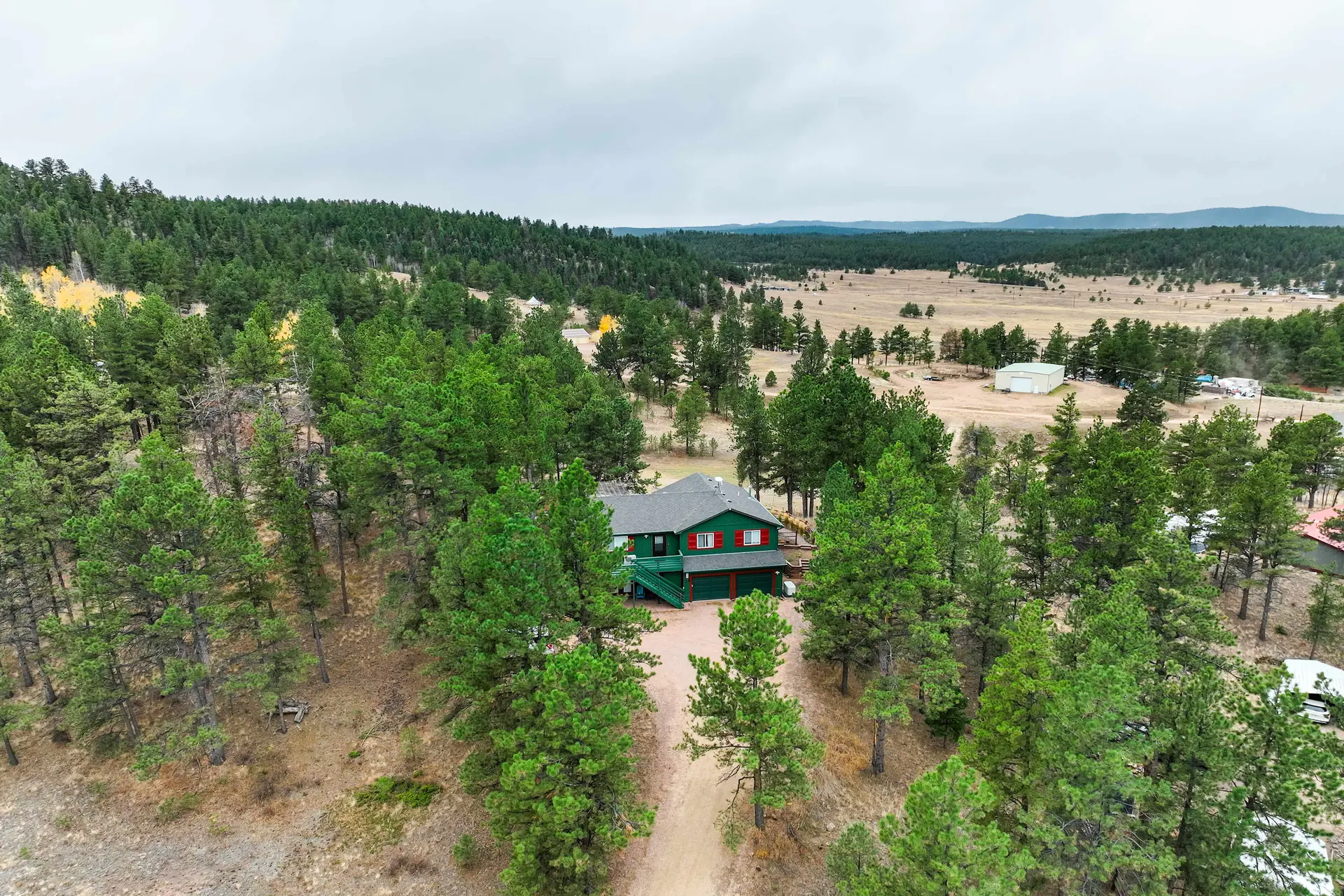 Aerial view of a green house in a dense pine forest, with a dry valley and mountains under a grey sk