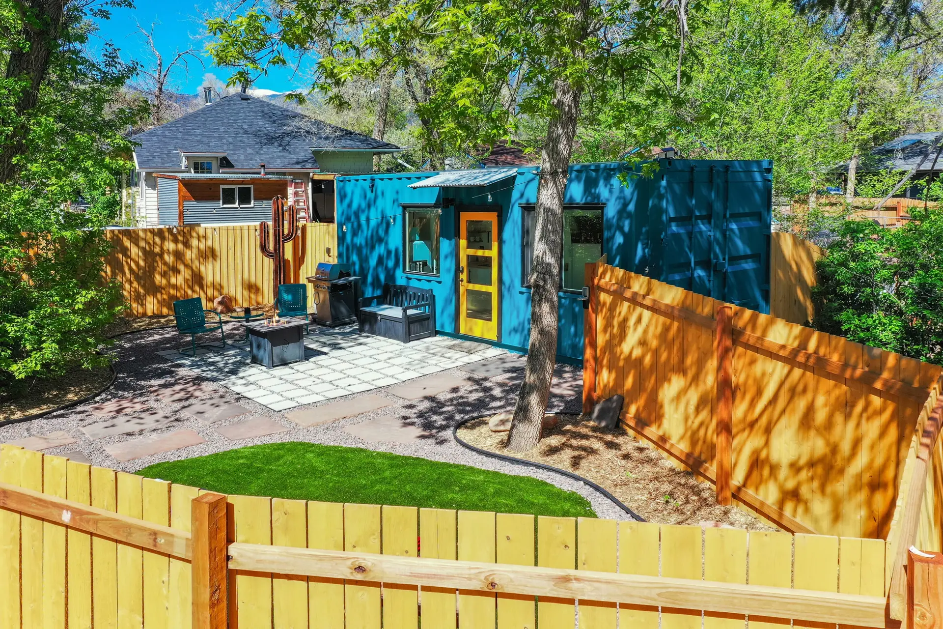 Bright blue container home with yellow door, patio, and landscaped yard with wooden fence under a su
