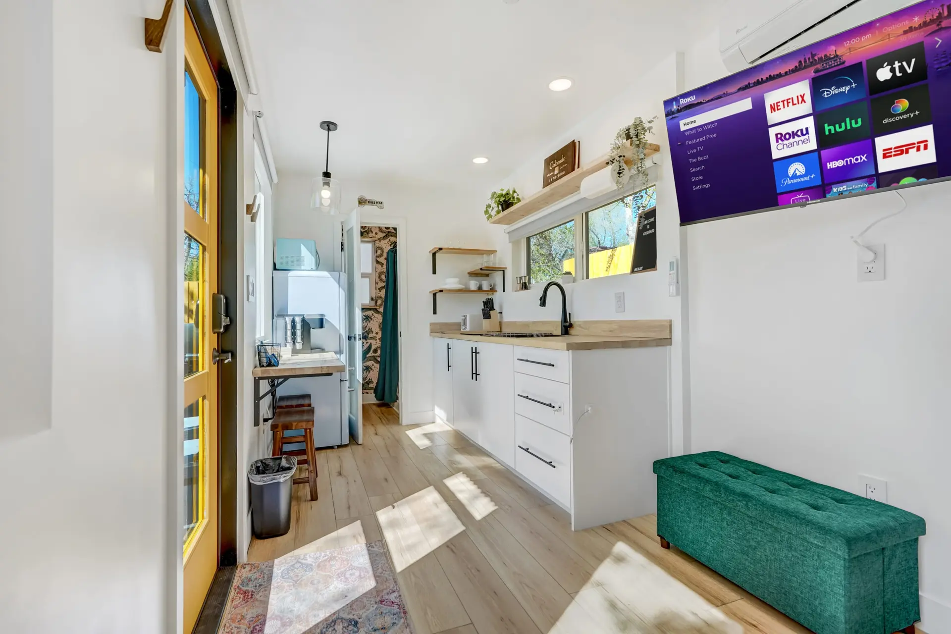 A bright, modern tiny home interior with a kitchen, a large TV showing streaming apps, and wood floo