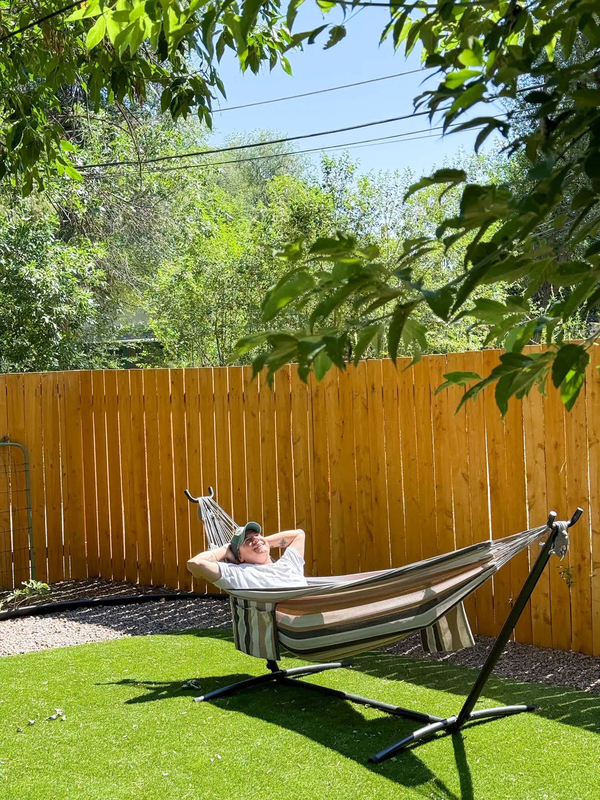 A person relaxes in a striped hammock on artificial turf, against a wooden fence and green trees.