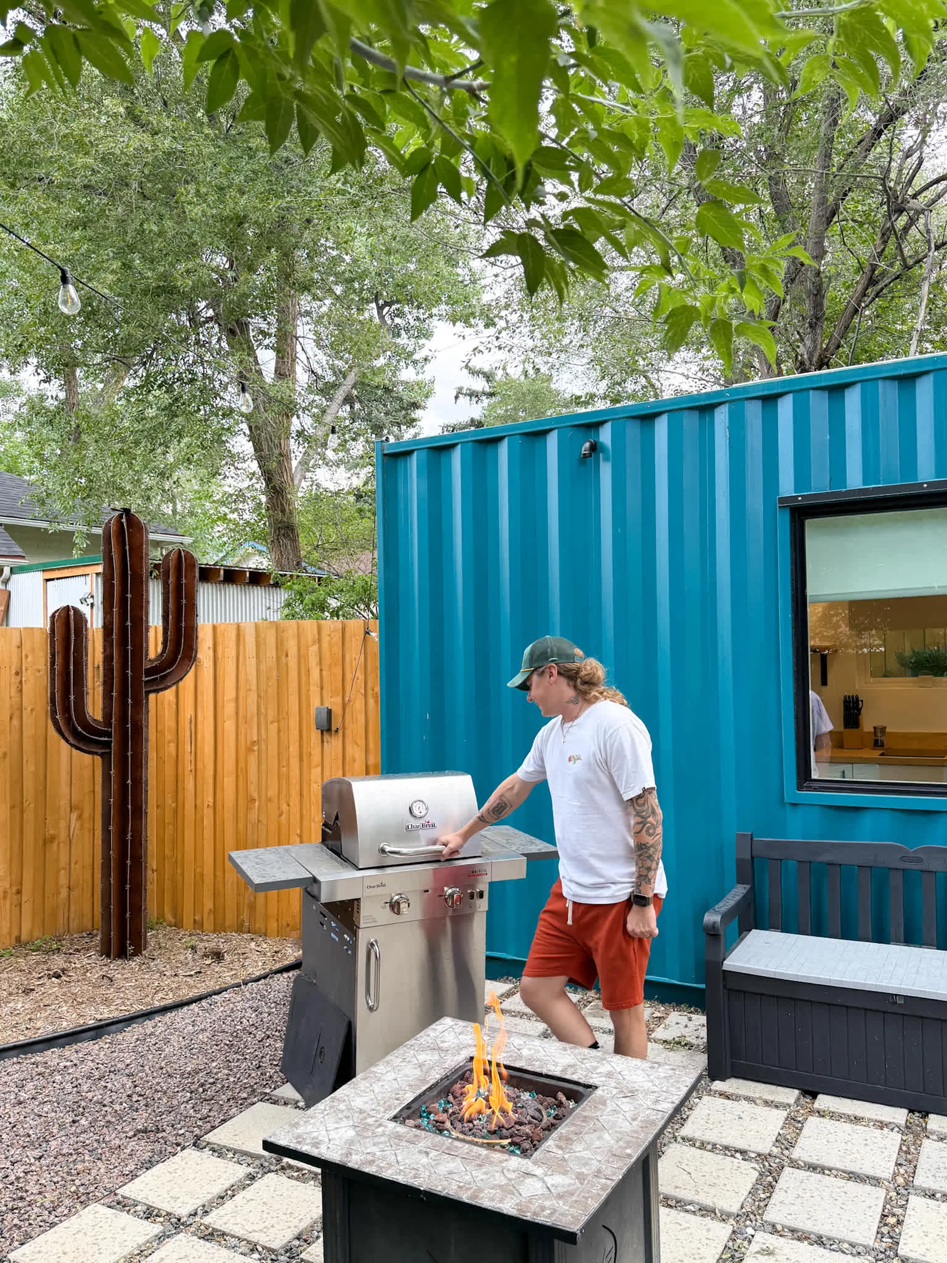 Man grills in a backyard featuring a teal shipping container home, fire pit, and metal cactus.