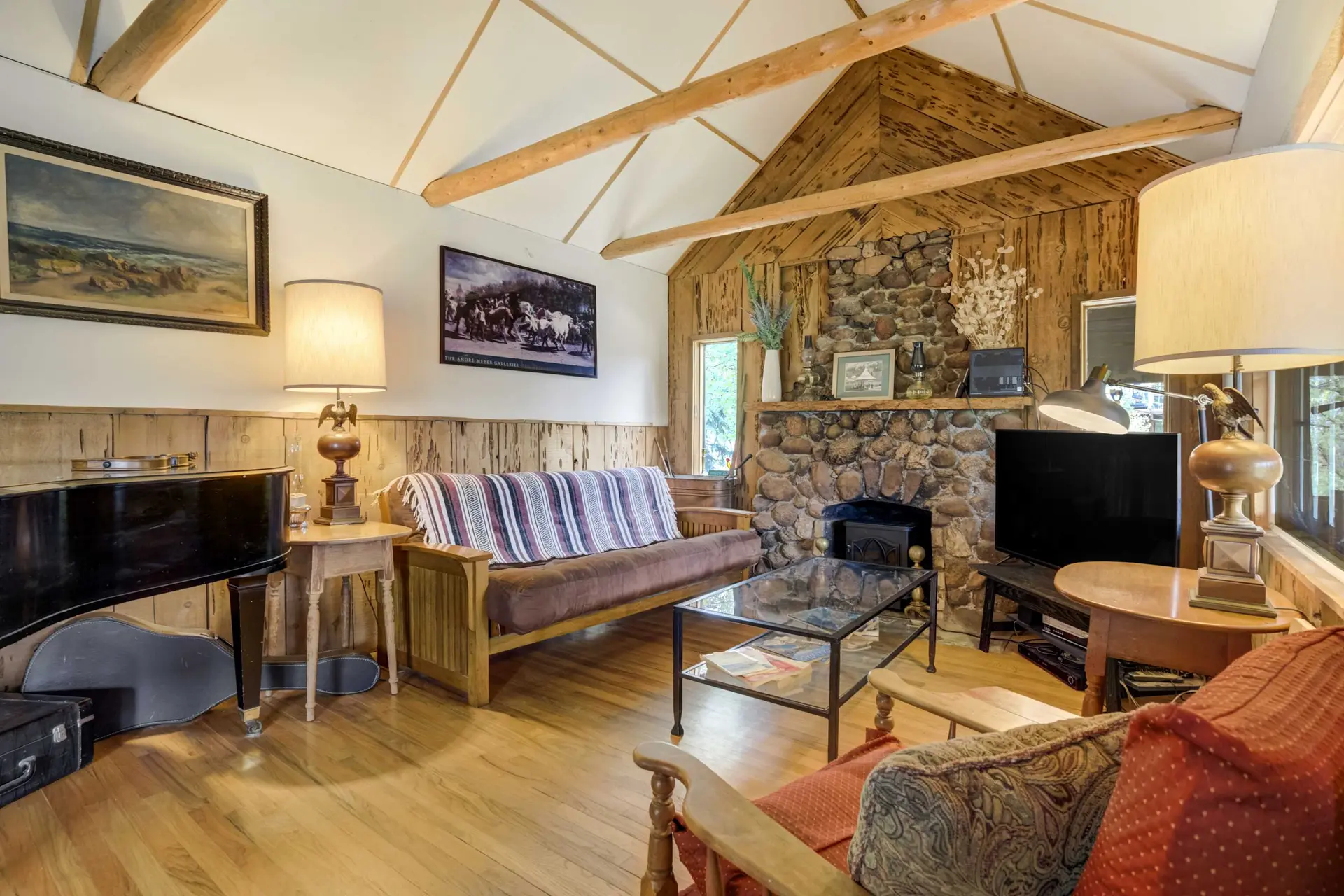 Rustic living room with stone fireplace, wood beams, futon, piano, and glass coffee table.