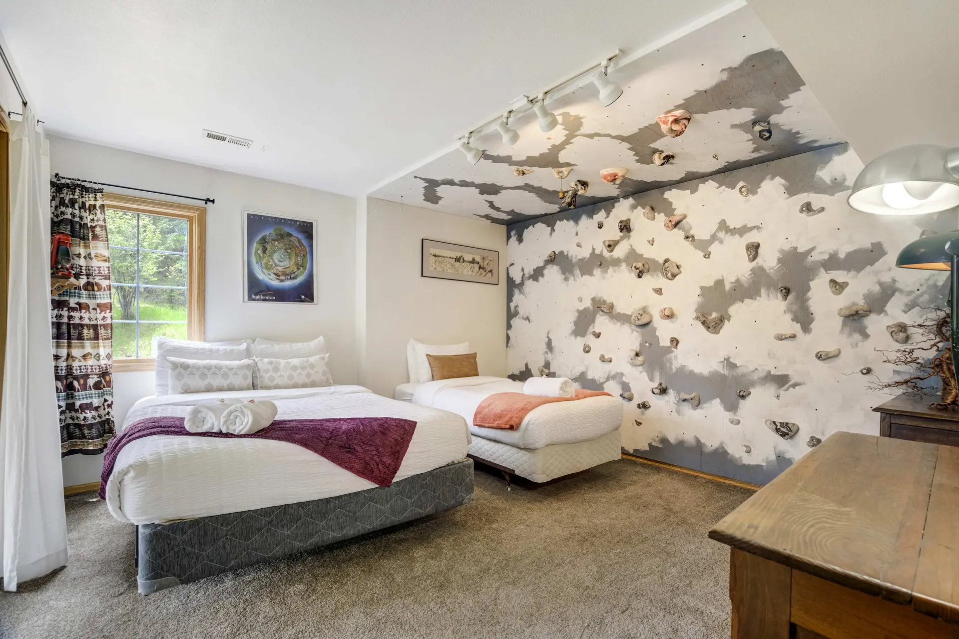 A bedroom featuring two beds, a climbing wall with holds, a window with animal-themed curtains, and 