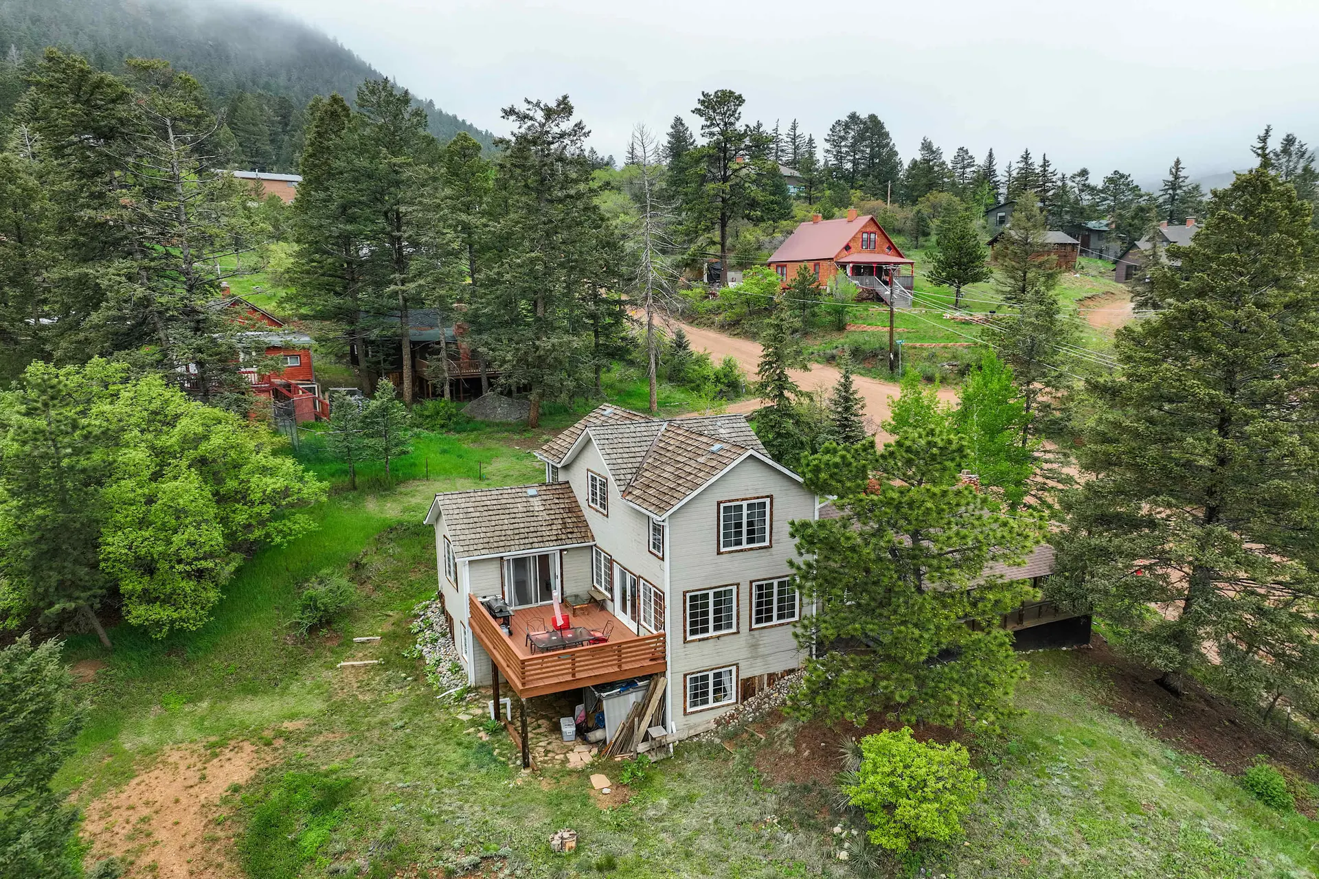 Aerial view of a light-colored house with a large deck in a green, forested mountainous area.