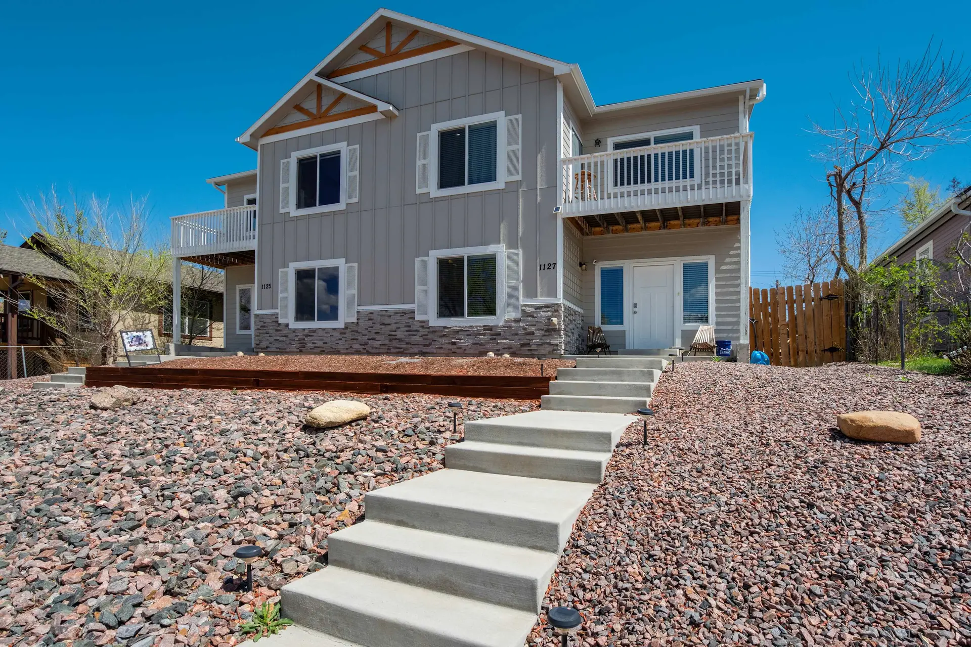 A modern gray two-story house with concrete stairs, gravel landscaping, and balconies under a clear 