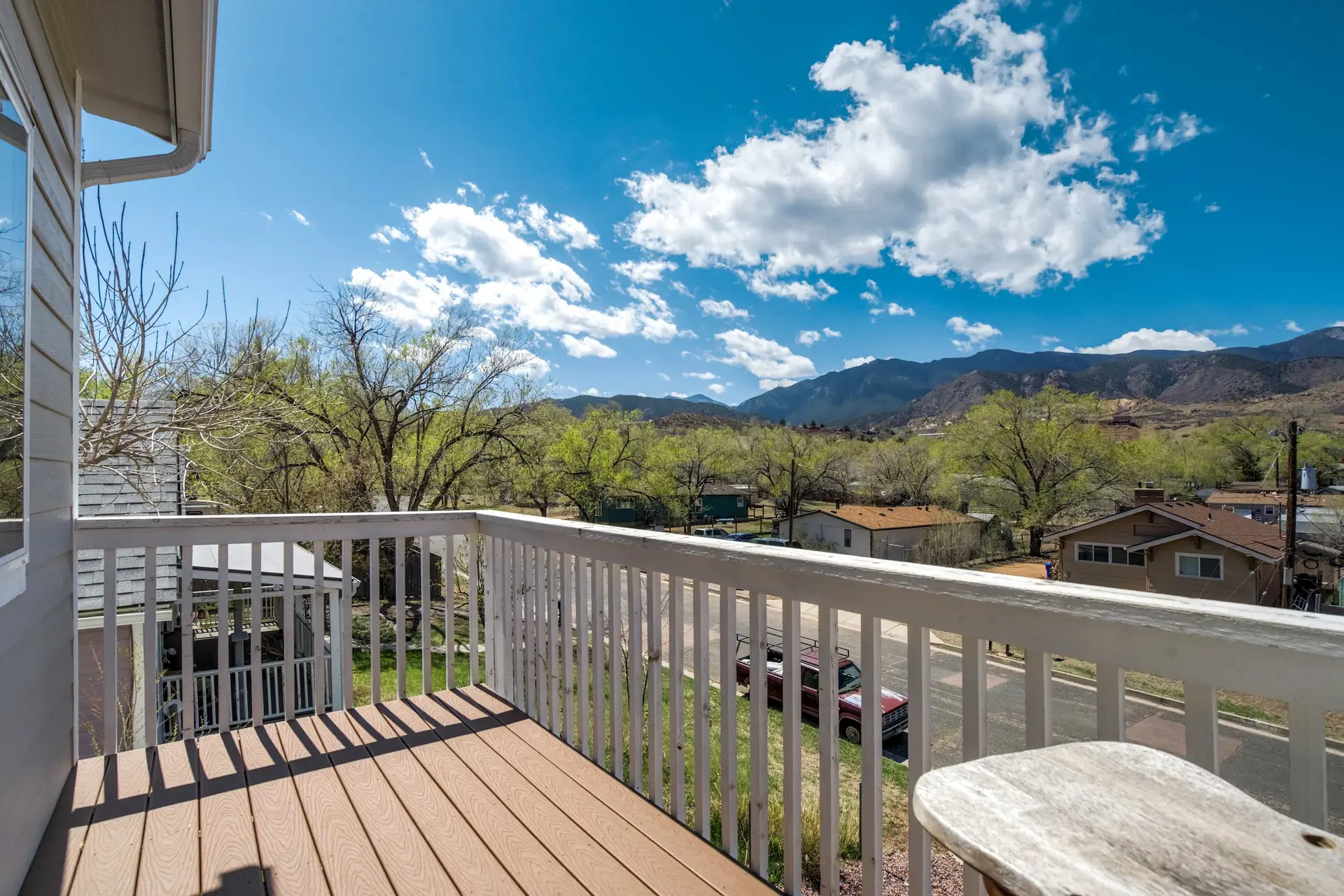 Sunny view from a balcony: wooden deck, white railing, houses, trees, mountains, and a blue sky.