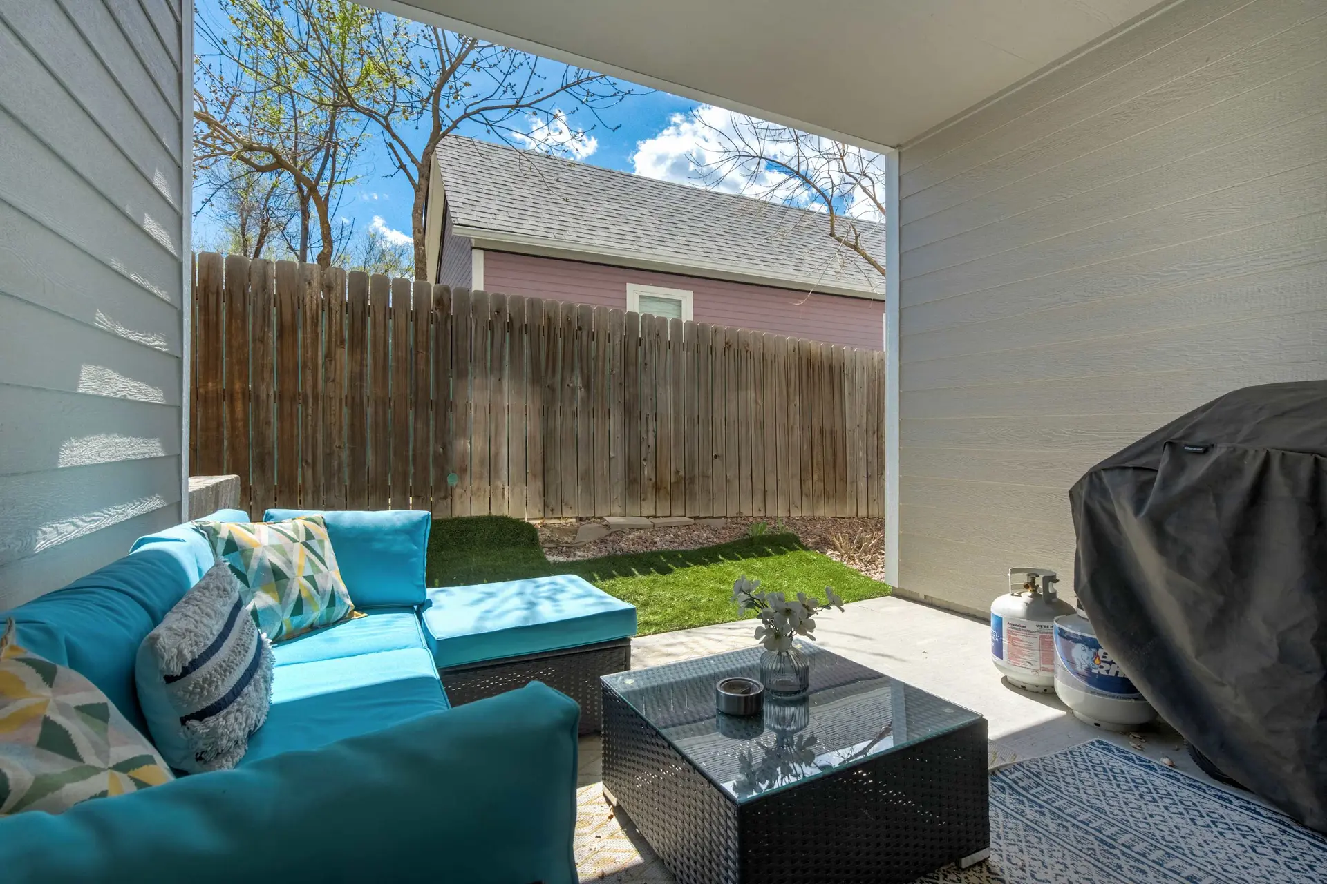 A sunny outdoor patio with a blue sofa, coffee table, covered grill, propane tanks, and a wooden fen