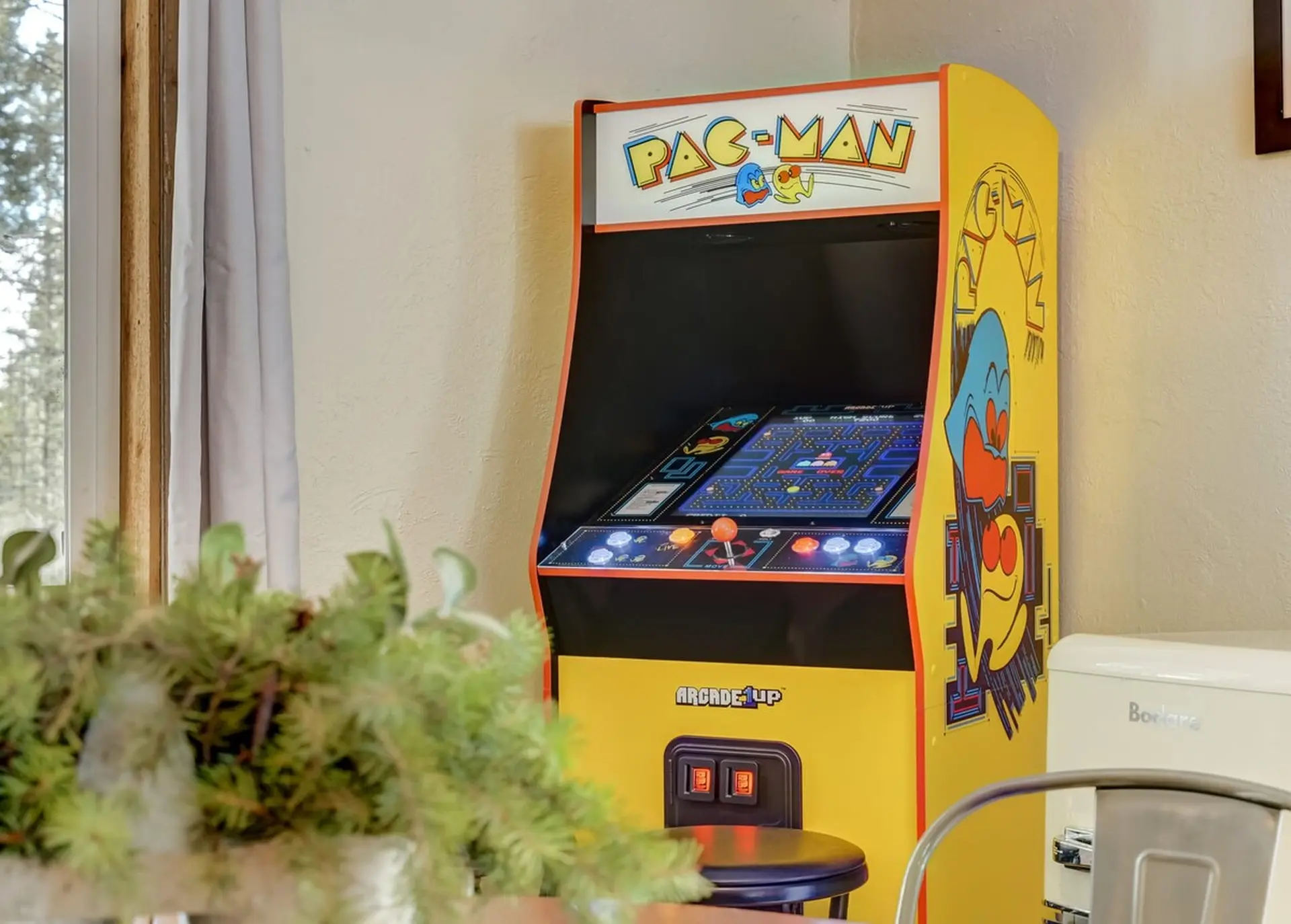 An Arcade1Up Pac-Man machine with a stool, a plant in front, and a window to the left.