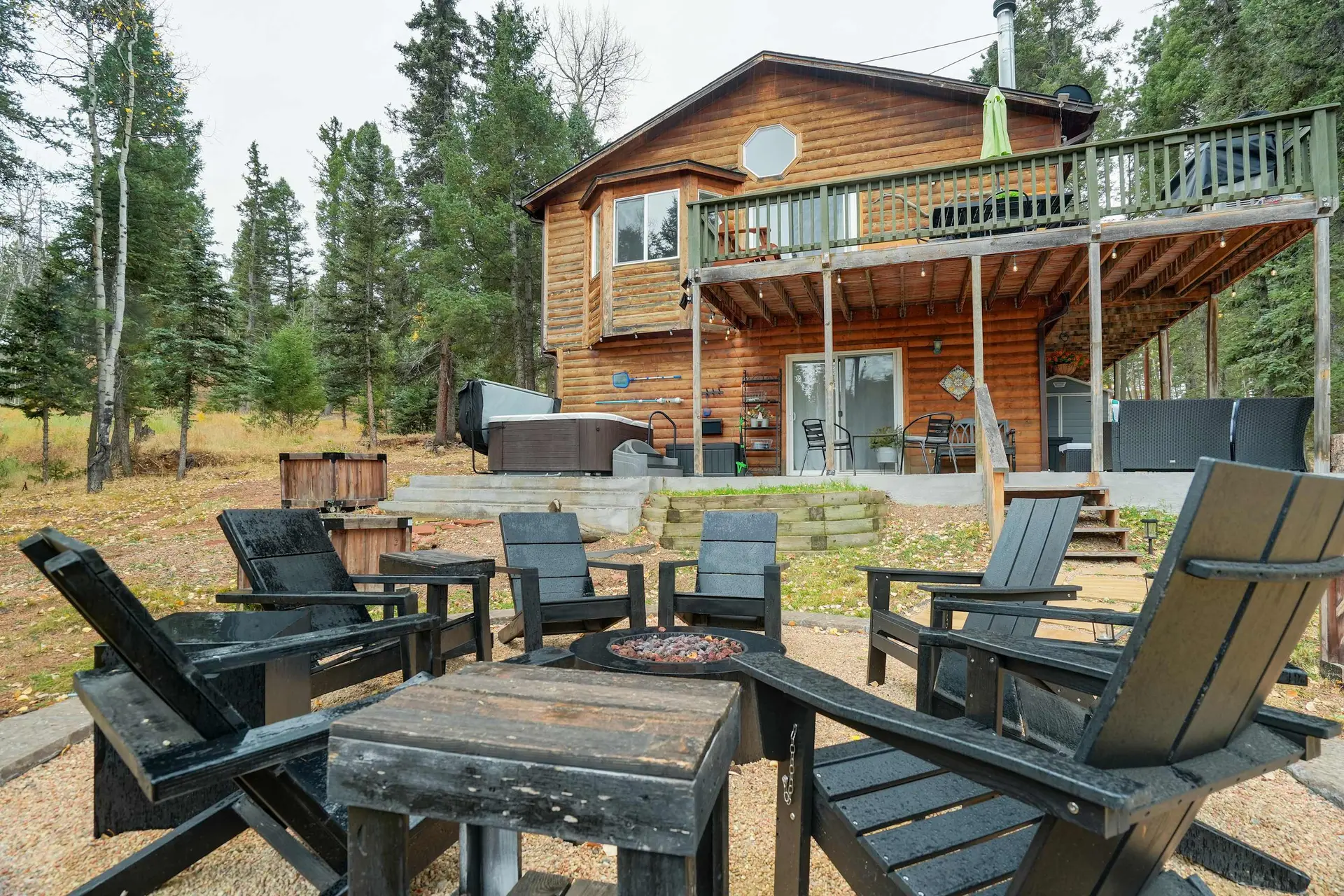 A log cabin house with a deck, hot tub, and a fire pit area with Adirondack chairs, surrounded by tr