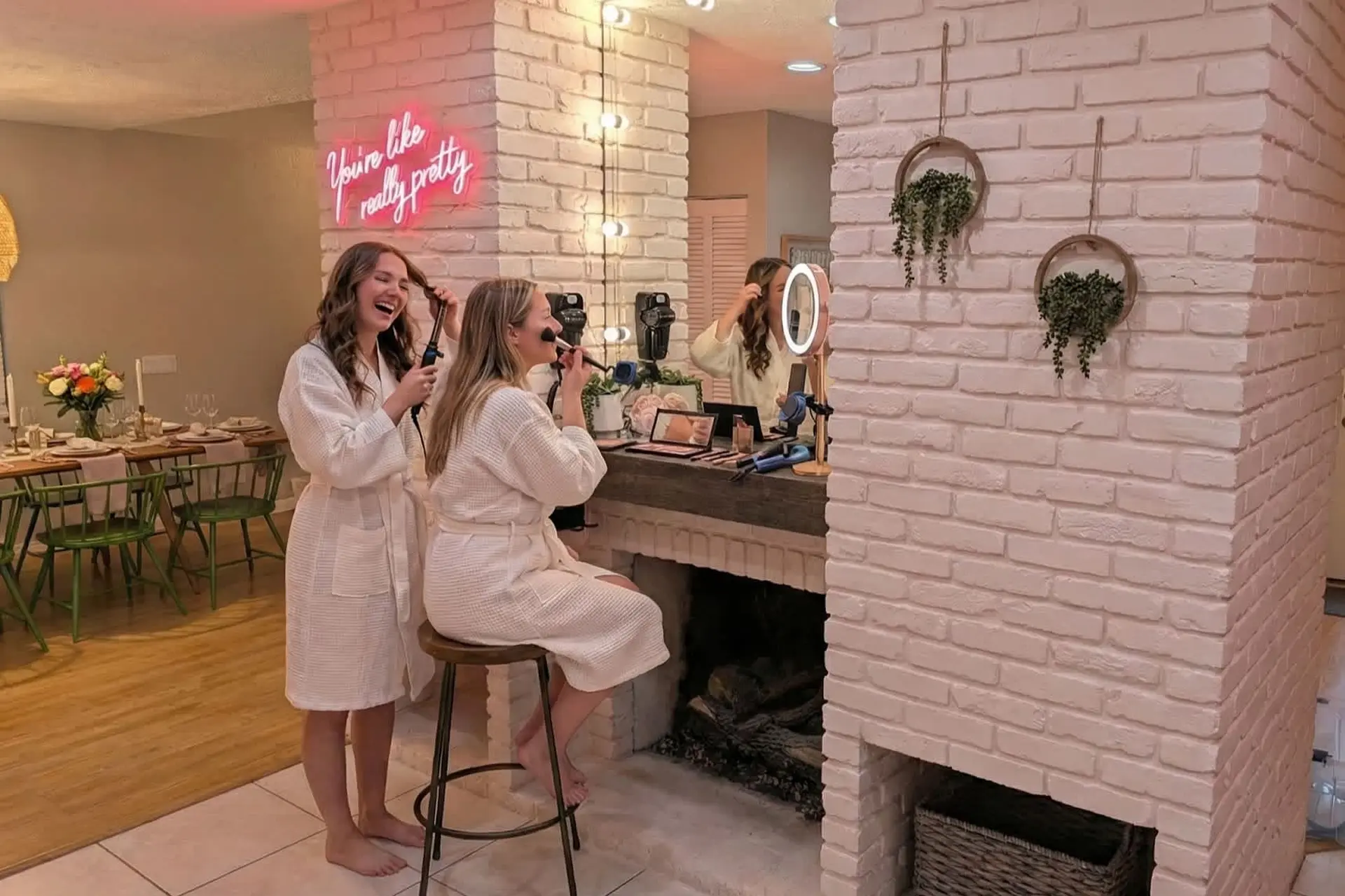 Two women in robes apply makeup in front of a fireplace, one with a curling iron, the other with a m