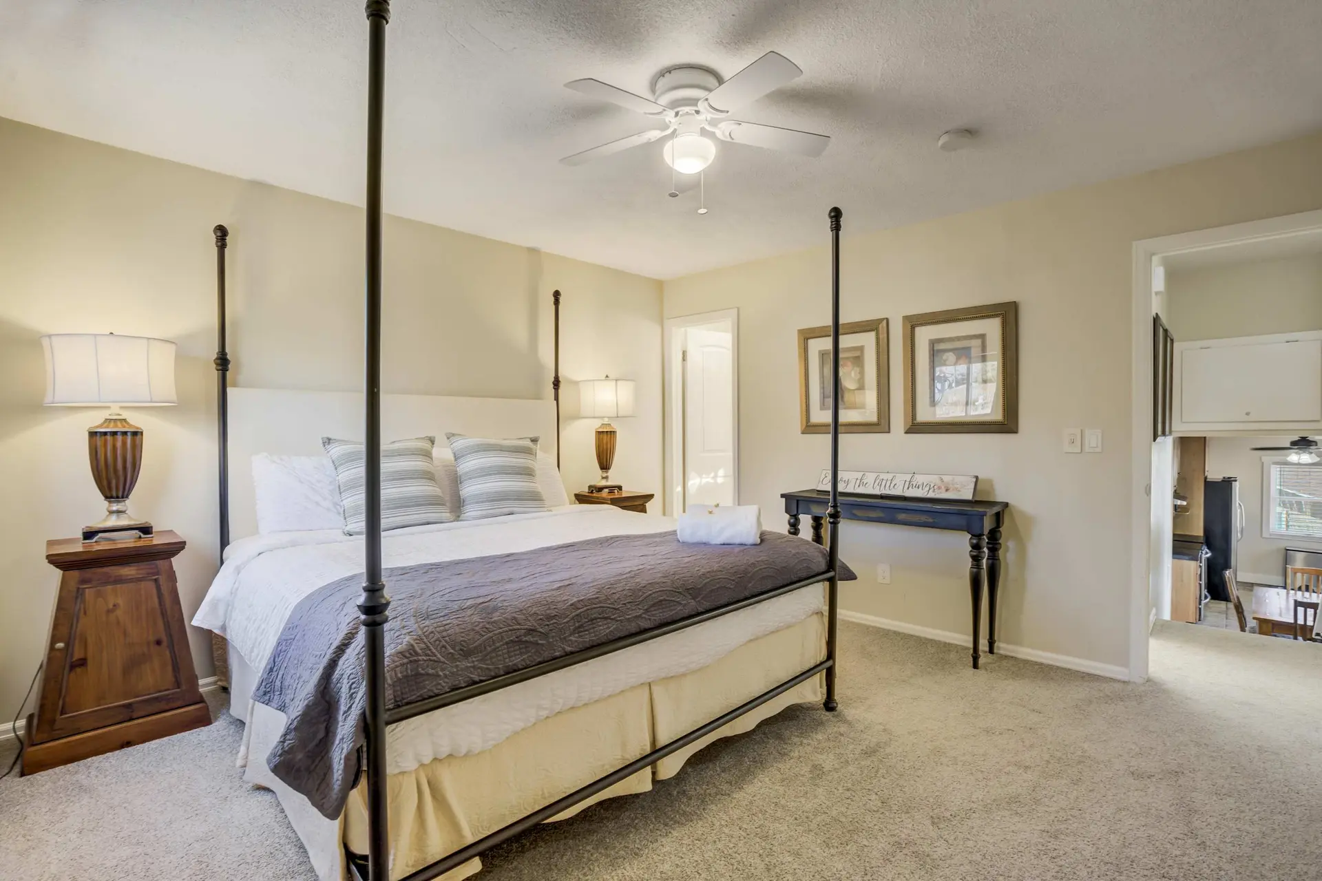A bright bedroom with a four-poster bed, lamps, ceiling fan, wall art, and a doorway.