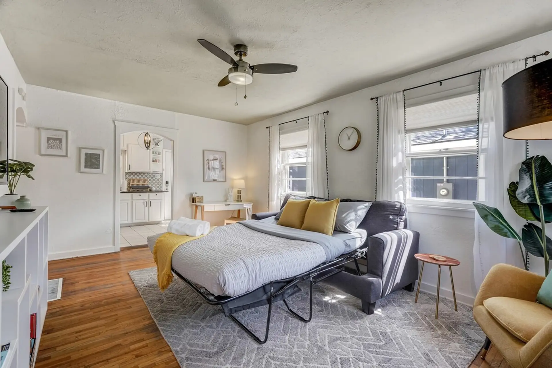 Well-lit living space with a grey pull-out sofa bed, a kitchen visible through an archway, and white