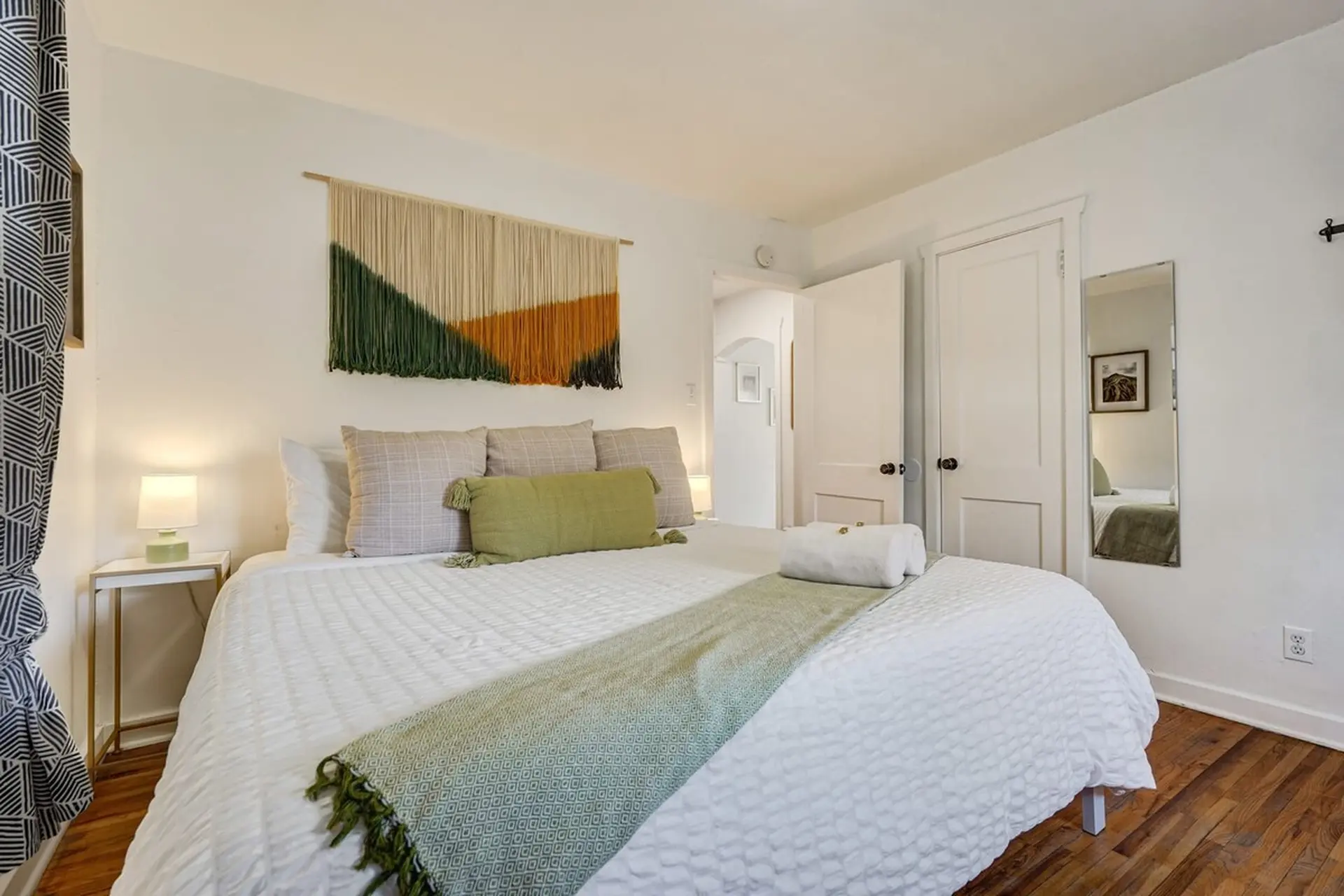 A cozy bedroom with a large bed, white bedding, green throw, and a colorful wall tapestry.