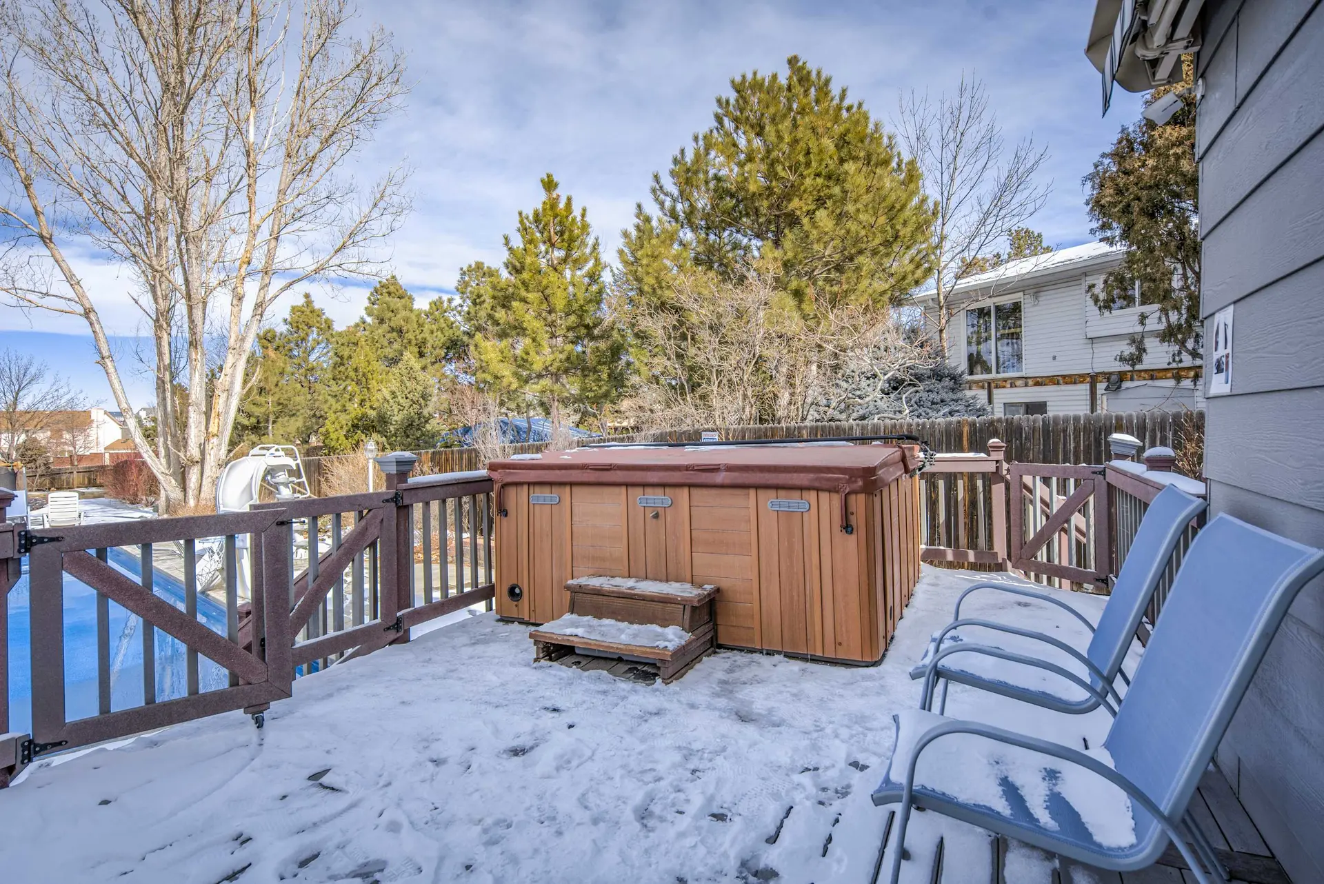 A wooden deck with a hot tub and chairs is covered in snow.