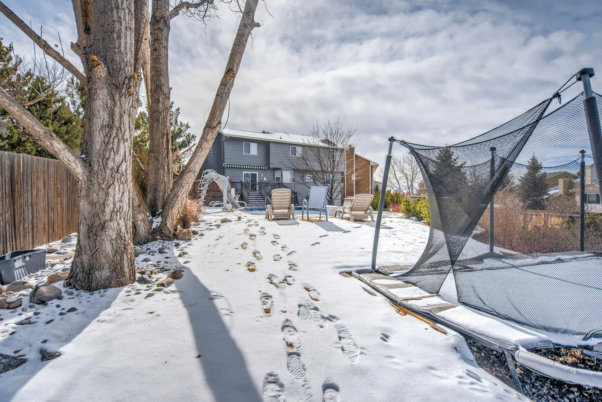 A snowy backyard with footprints leading to a trampoline and lounge chairs, with a gray house in the
